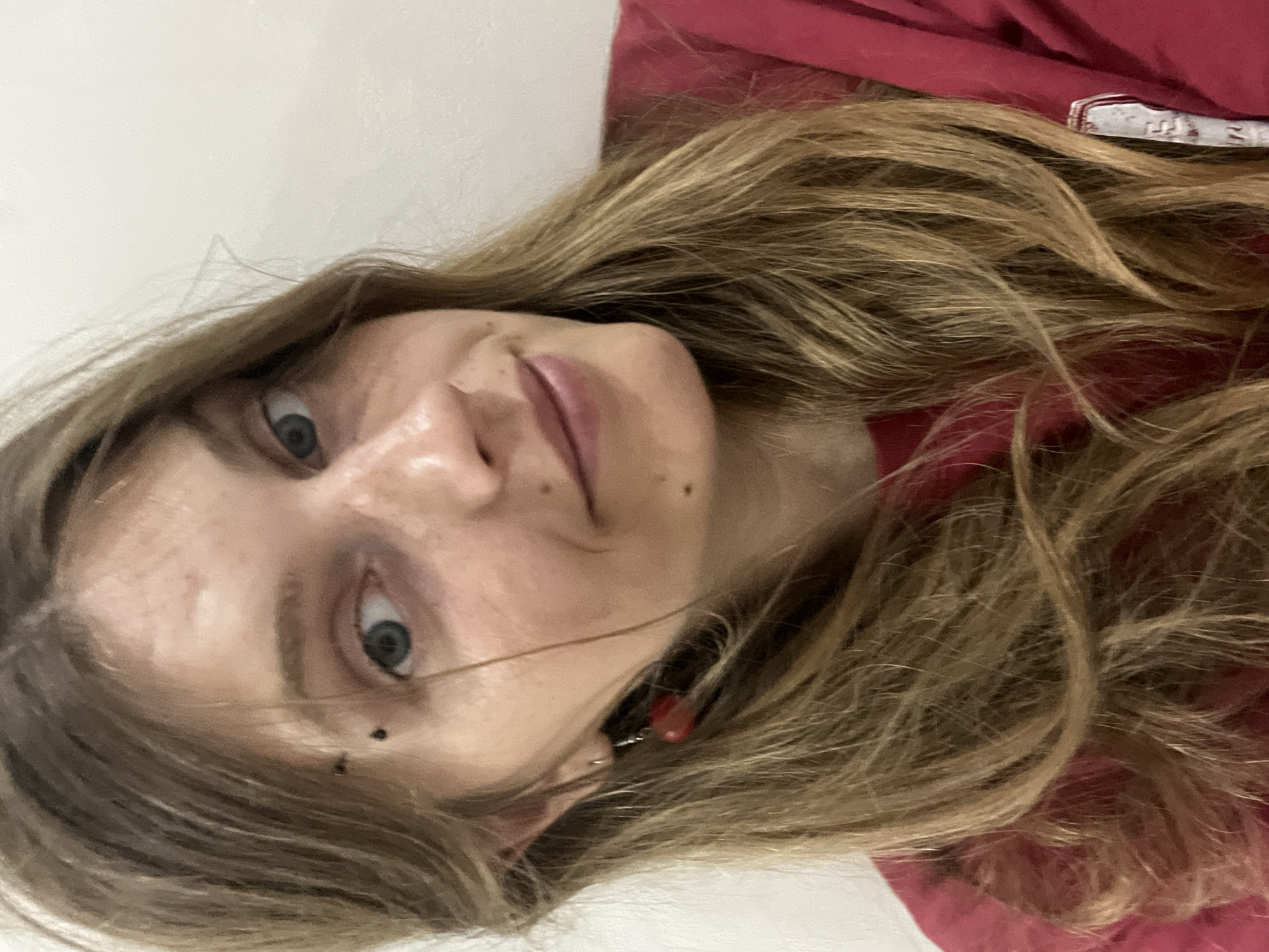 A close-up, head-and-shoulders selfie of a young woman with blonde, shoulder-length hair. She is wearing a red shirt and has a small black piercing near her left eye. She is also wearing a red, dangle earring in her left ear. Her eyes are blue and she has freckles on her face. The background is a plain white wall. The image was taken indoors, likely in a home. The location of Сеніцкі сельскі Савет, Belarus is not visible in the image.