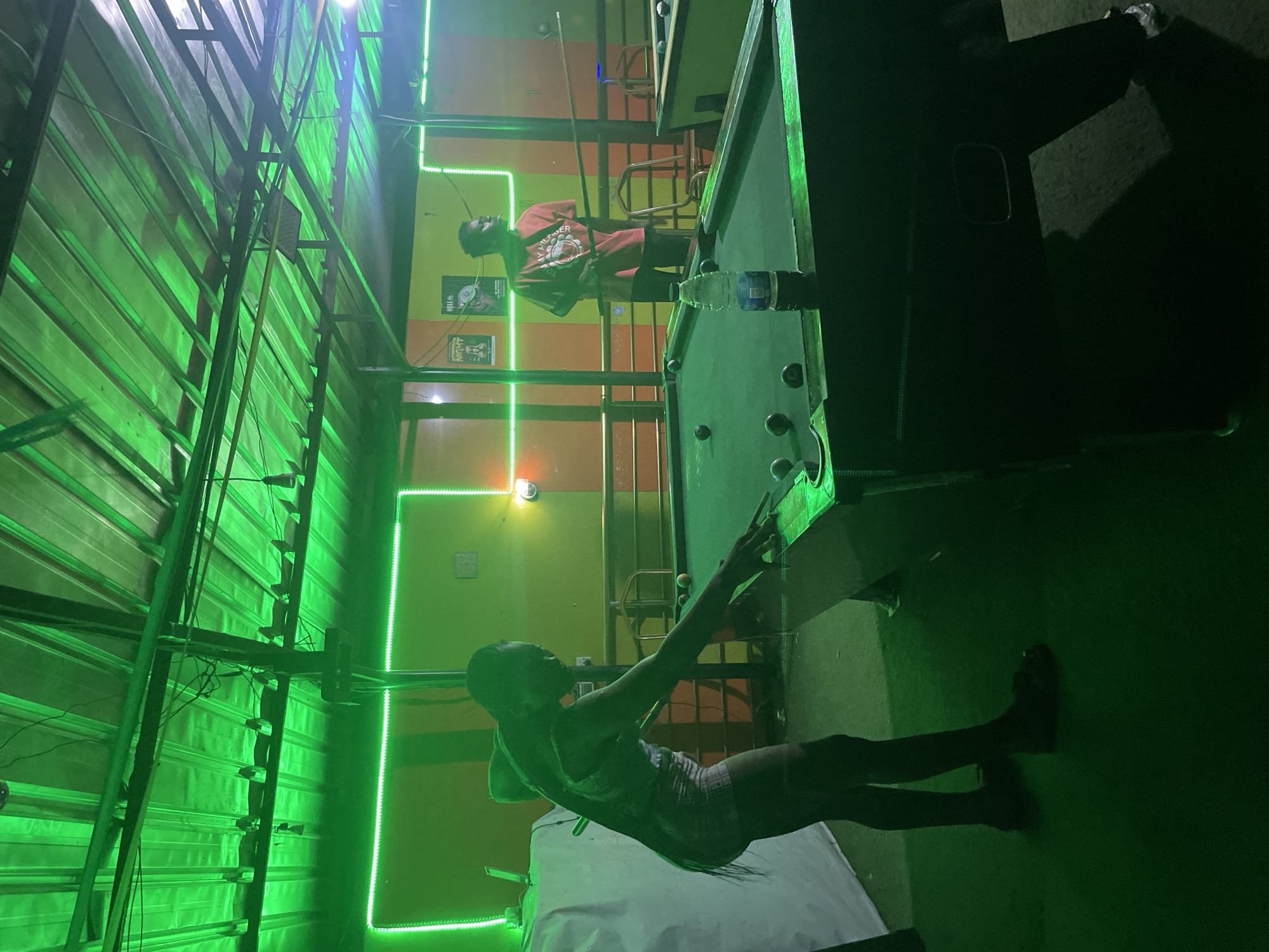 The interior of a dimly lit pool hall in Ado, Nigeria is shown. The lighting is primarily green, emanating from neon strips. Two men are present: one leans over a pool table, cue in hand, while the other stands near a second table in the background. A water bottle sits on the pool table. The room's structure is metallic, with corrugated roofing visible. Two promotional posters hang on a green and orange wall.