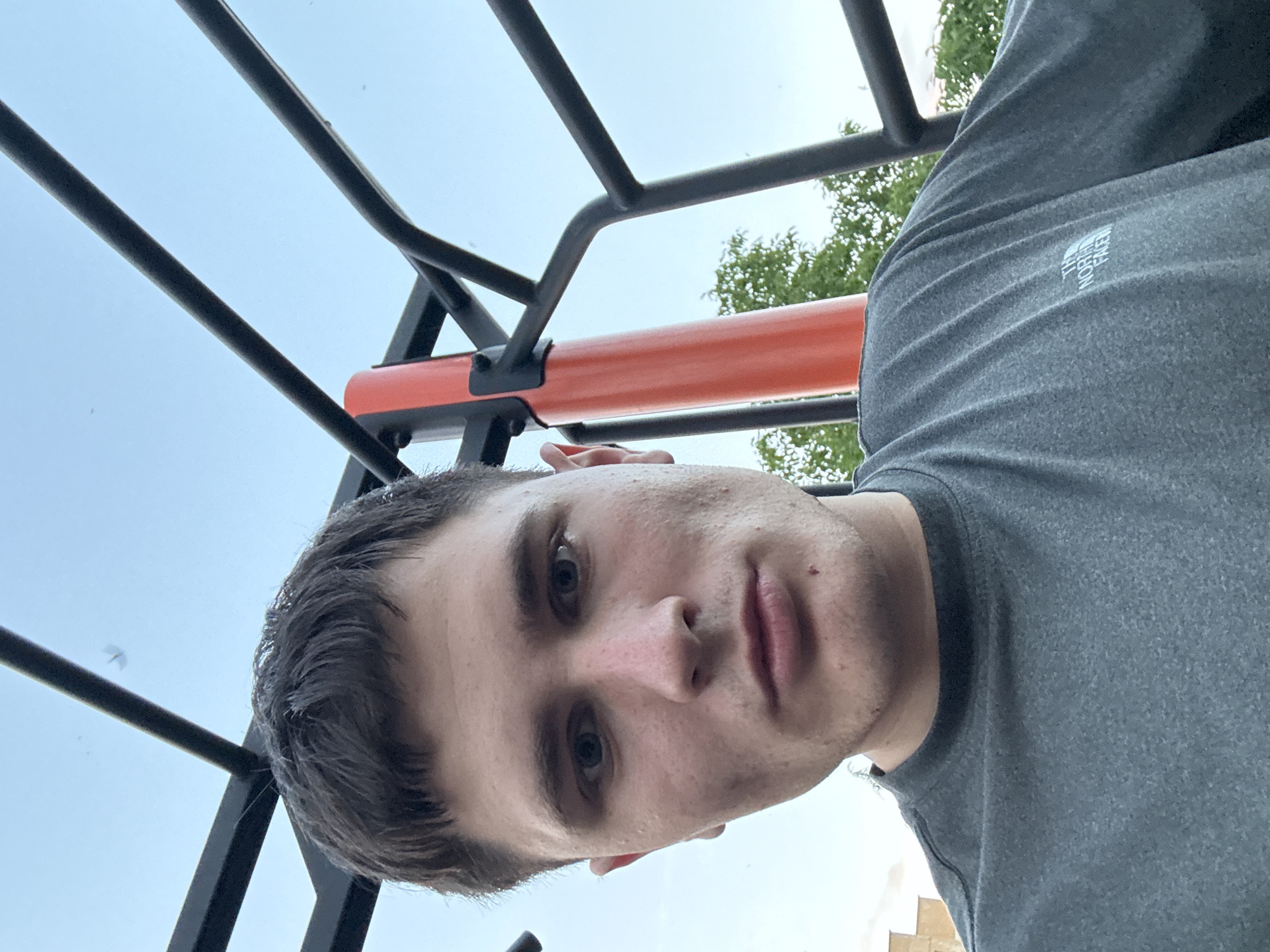 A young man with short, dark hair is pictured from the chest up, looking directly at the camera. He is wearing a dark grey t-shirt with a small white logo on the left side of his chest. The background consists of a blue sky, a black metal structure resembling monkey bars or a playground frame, and a section of an orange pole. Green foliage is visible behind the metal structure, suggesting an outdoor setting. The image appears to be a selfie, taken from a low angle.