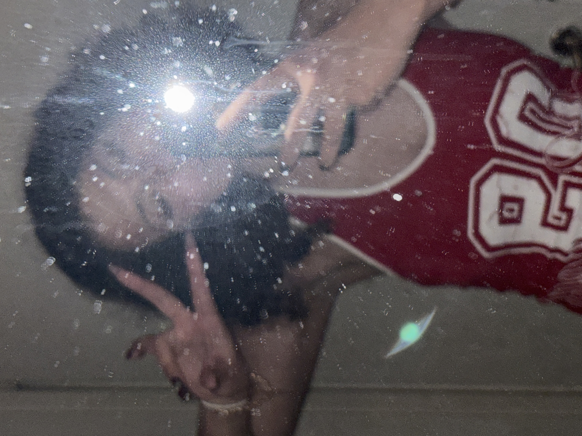 The image depicts a selfie taken in a mirror. The person in the mirror is wearing a red jersey with the number 80. The person is making a peace sign with their left hand and holding the phone with their right hand. The mirror is dirty. The flash of the camera creates a large glare.
There is no evidence to indicate the location is Milan, Italy.