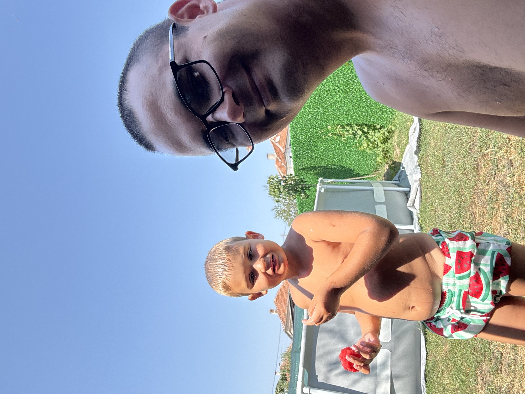 This media file captures a playful outdoor moment on a sunny day. Two individuals are present: an adult wearing glasses and a young child. The adult's face is partially visible, looking downwards. The child, who appears to be a boy, is in the foreground and is shirtless, wearing swim trunks with a watermelon pattern. He is holding a red object in his hand.
The scene appears to be a backyard or outdoor recreational area. In the background, there is green grass, a white fence, and a part of an inflatable pool. Some trees and buildings with red roofs are also visible, suggesting a residential setting. The clear blue sky indicates fair weather, and the bright sunlight suggests it is daytime.
The overall mood seems to be one of fun and relaxation, likely during warm weather, perhaps a summer day. The child's expression is one of engagement and possibly enjoyment. There is no visible text in the media file. The context provided places this scene in Svilengrad, Bulgaria.
