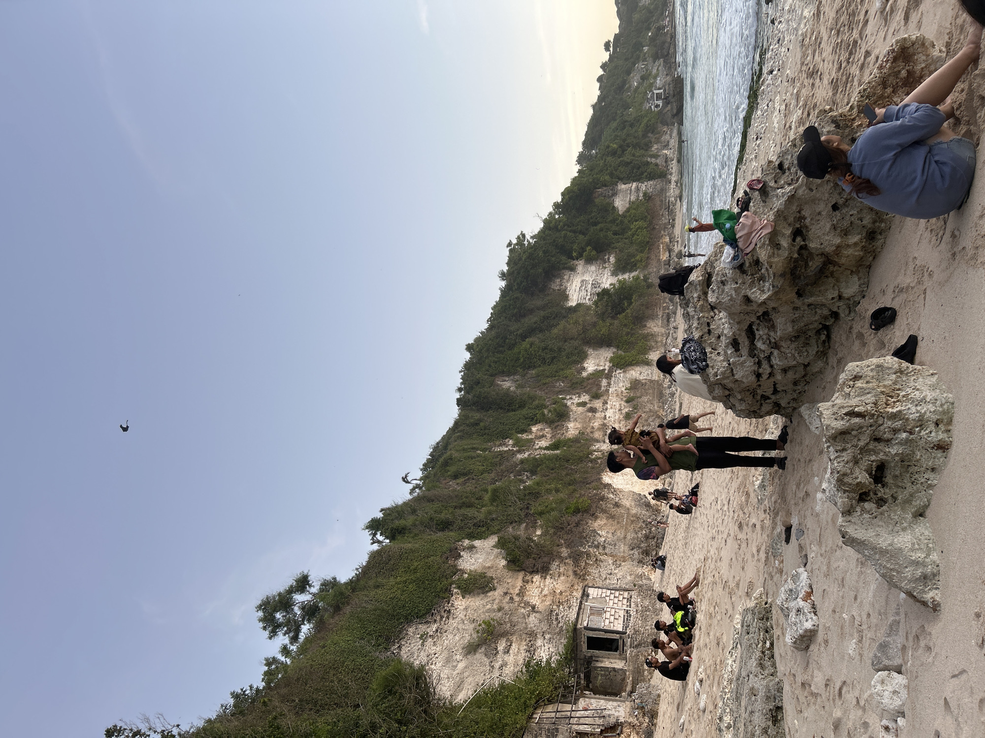 The image captures a scenic view of a beach with a cliff face covered in lush green vegetation in the background, likely located in Kuta Selatan, Indonesia. The sky is a clear, pale blue with a single dark bird in flight. On the sandy beach, several groups of people are present. In the mid-ground, a man is holding a child, with other individuals sitting or standing nearby. Further down the beach, more people are scattered, some relaxing on the sand, others near the water. The foreground features large rock formations and the sandy shore. The ocean is visible on the right side of the image, with gentle waves rolling in. The cliff face displays layers of rock and dense foliage, with some dilapidated structures visible at its base. The overall scene suggests a relaxed atmosphere on a tropical beach.