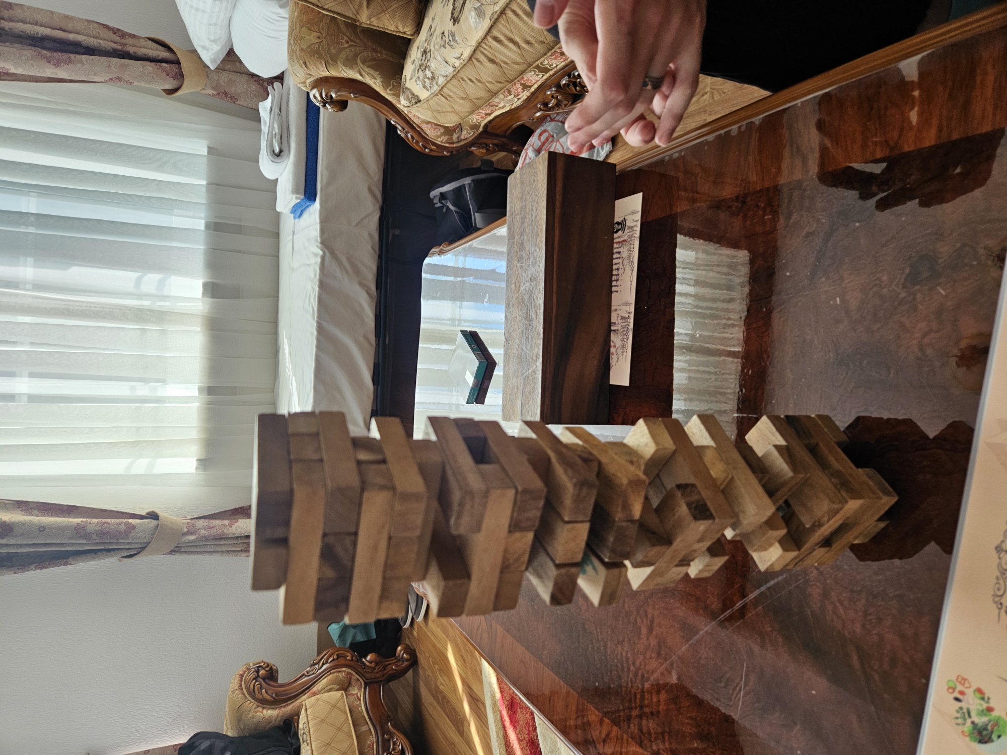 The image shows a table with a wooden block puzzle on it. The puzzle is made of interlocking wooden pieces. A person's hands are visible over the puzzle, suggesting they are either playing with it or have just finished. The table is made of polished wood and reflects the light. In the background, there is a bed with white linens and a decorative armchair. There are also curtains and blinds on a window. The scene appears to be indoors, possibly in a hotel room in Krasnaya Polyana, Russia, given the mention of the location. The lighting suggests it could be daytime. There is some text on a piece of paper next to the puzzle, but it is not legible. The overall impression is one of leisure or a moment of quiet activity.