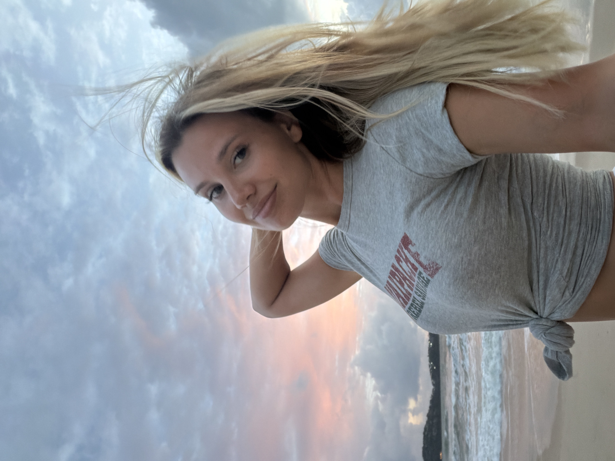 An outdoor, eye-level, medium shot captured on a sandy beach in Choeng Thale, Thailand, shows a woman with blonde hair in a grey t-shirt tied at her waist. She is looking at the camera with her left arm raised behind her head, and her hair is blowing in the wind. The ocean is to her left, with waves breaking on the shore. The sky is filled with clouds in shades of grey and pink, indicating it is either sunrise or sunset. The background features a distant shoreline with trees and lights.
