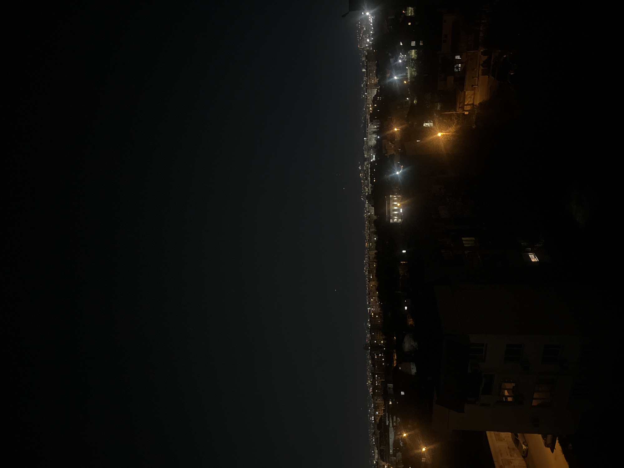 The image shows a night view of Sofia, Bulgaria. The right side of the image depicts buildings illuminated by streetlights and lights from within. The left side of the image shows a dark night sky with a few faint stars. The cityscape appears to stretch into the distance, marked by scattered lights. The specific location cannot be pinpointed without further contextual information.