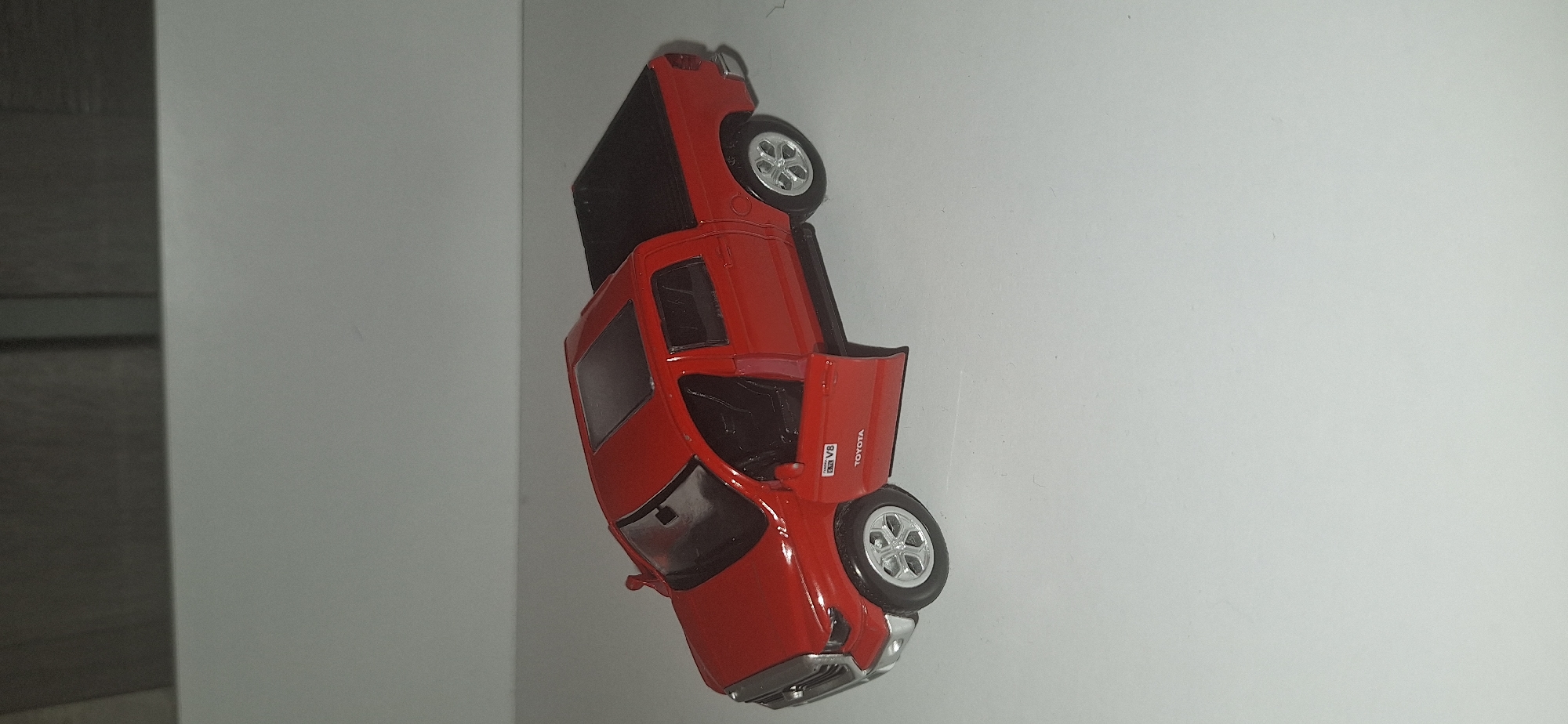 A red toy Toyota Tundra pickup truck is displayed on a white surface. The truck has its driver's side door open, revealing black interior seats. The truck bed is black and covered. There is a sunroof on the cabin's roof. The truck has silver rims on its wheels. The background is out of focus, showing a grey, textured surface. There is no indication of the location being in Bila Tserkva, Ukraine.
