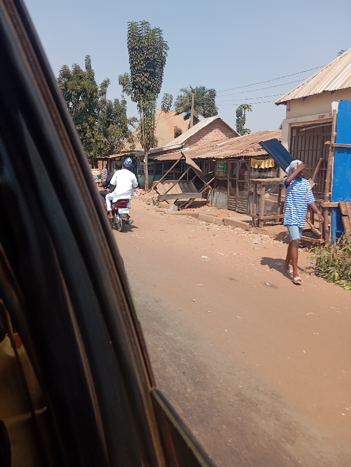 The image depicts a street scene in Kadarko, Nigeria, captured from the interior of a vehicle, looking out a dark-framed window or doorway on the left. The scene is illuminated by bright daylight under a clear, pale blue sky.

A reddish-brown, unpaved road occupies the center and lower right of the frame, showing signs of dust and scattered debris. In the mid-ground, a male individual on a dark-colored motorcycle, wearing a white long-sleeved garment and a light-colored cap, rides away from the viewer. Ahead and to the right of the motorcycle, another male individual walks leftward across the frame, carrying a dark, rectangular object, consistent with a corrugated metal sheet, balanced on his head and steadied by his left hand. He wears a blue and white striped short-sleeved shirt and light-colored shorts or trousers.

Along the right side of the road, a series of single-story buildings line the street. Many feature corrugated metal roofs, some with brick or concrete walls, and others appear to be constructed from wood or makeshift materials, serving as shops or stalls. Some wooden structures in front of these buildings appear in varying states of repair. A notable sign on a building on the far right partially reads "GOOD T […]," adjacent to a temporary blue panel. Trees, including one tall, slender specimen with dense foliage at its apex, are visible behind the structures and in the background. Sparse ground vegetation is present along the roadside.