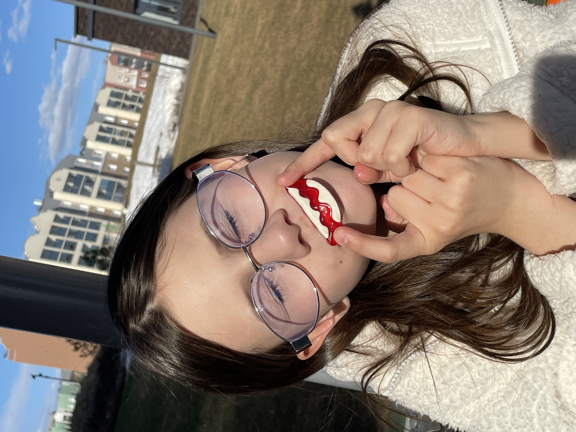 A young person with brown hair is posing outdoors on a sunny day, holding a half-moon shaped, white and red object (possibly candy or a theatrical prop) near their mouth, in front of a blurred background of multi-story residential buildings and snowy patches. The person is wearing glasses with light purple lenses and a white, fluffy jacket. The buildings in the background suggest the location is in the city of Pervomaysky, Russia.