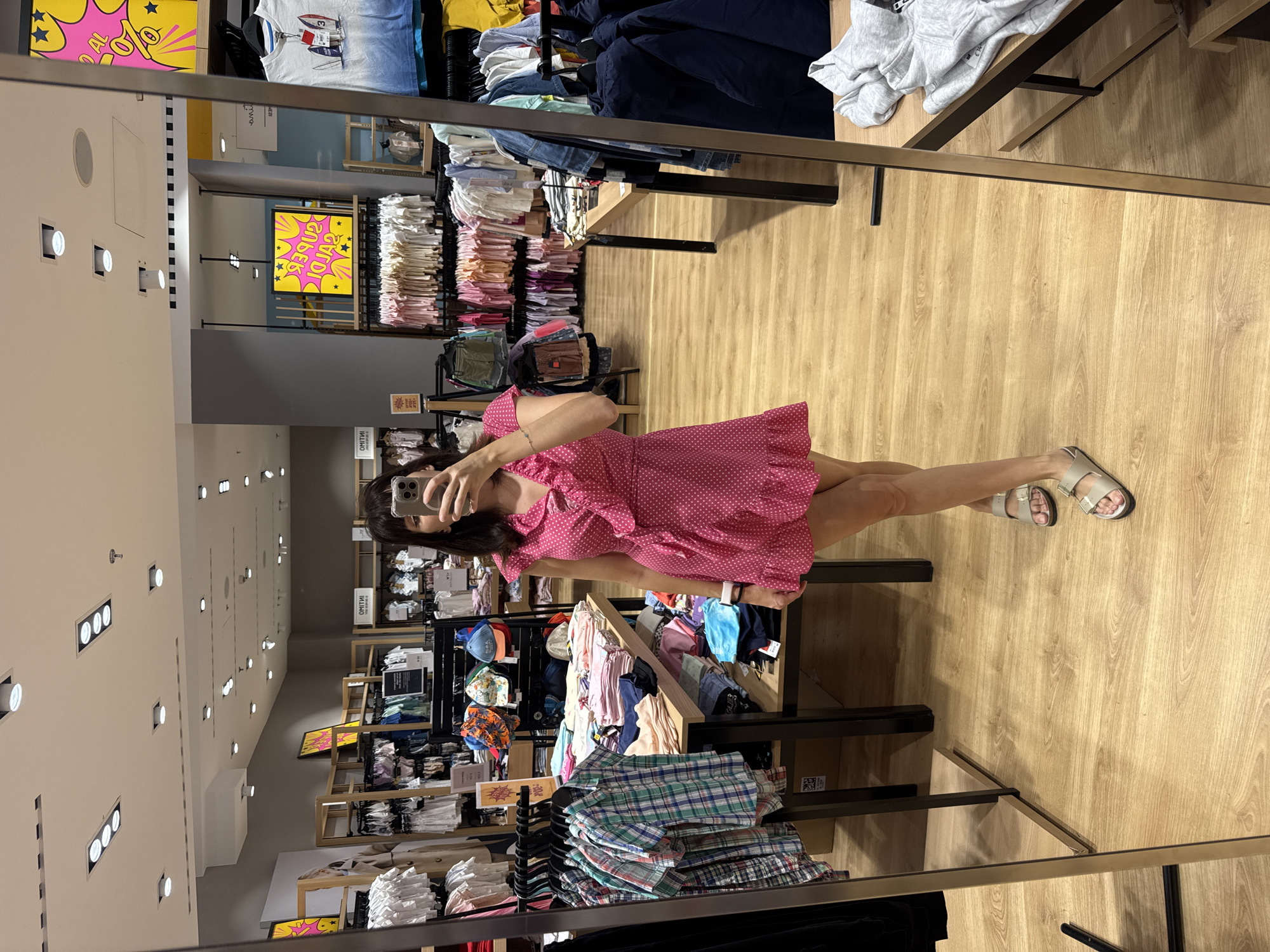 A person is standing in front of a mirror in a clothing store, taking a selfie. The person is wearing a pink polka dot dress and sandals. The store is filled with clothes on racks and shelves. The mirror reflects the store, including racks of clothing, displays, and other shoppers. The floor is made of wood. The store is located in the city of Carini, Italy.