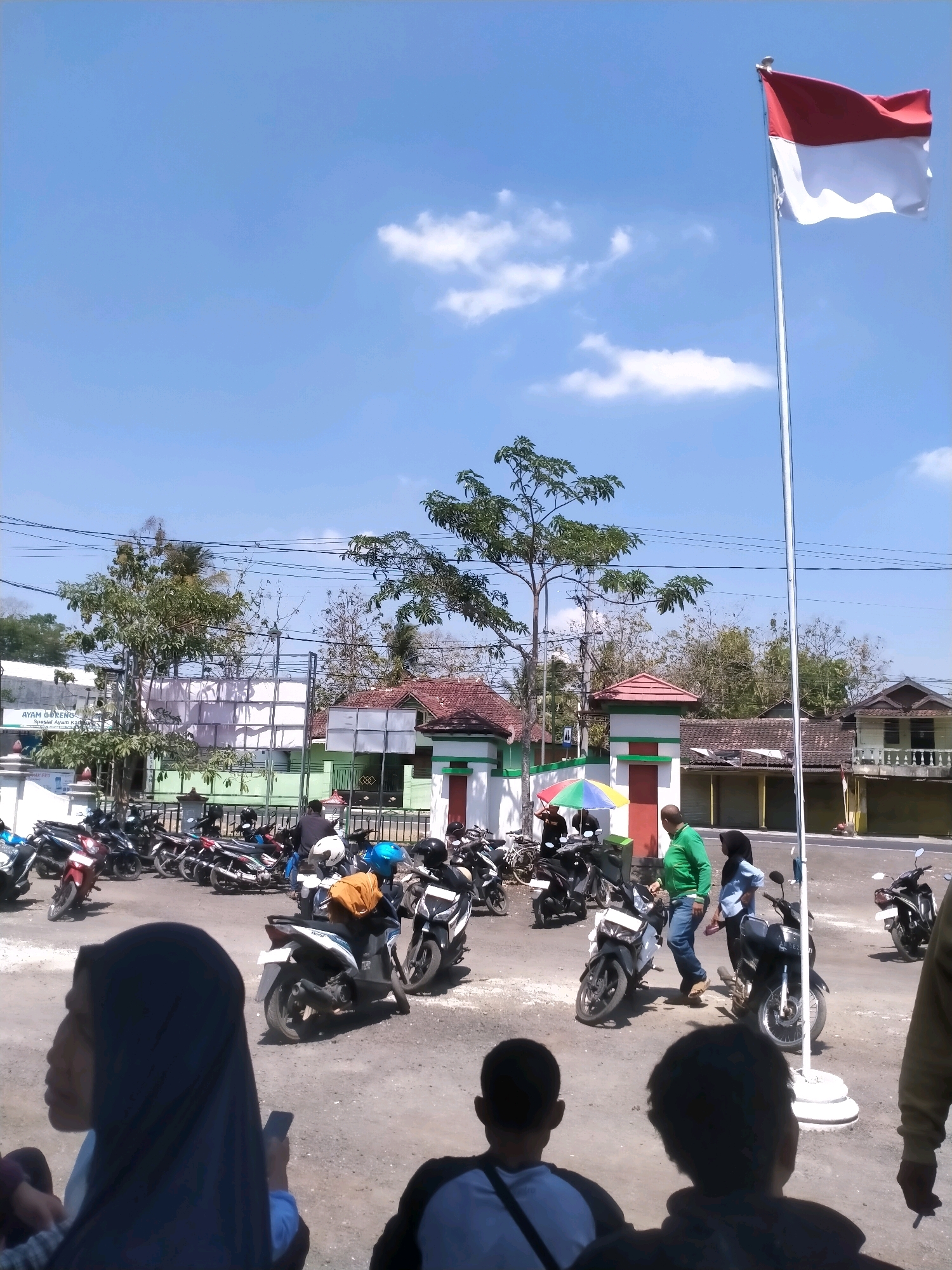 The image captures an outdoor scene in Playen, Indonesia, on a bright, sunny day with a clear blue sky dotted with a few white clouds. In the foreground, a red and white flag, likely the Indonesian flag, flutters on a tall flagpole, positioned to the right of the frame. The ground is a gravelly, dusty surface, with several parked motorcycles filling a significant portion of the area.
Several individuals are present in the scene. In the immediate foreground, facing away from the camera, are the backs of two people, one wearing a dark top and the other a dark jacket. To the left and slightly further back, a person wearing a dark blue hijab is looking towards the left, holding a mobile phone.
Further into the scene, a group of people are interacting near some parked motorcycles. One person, wearing a bright green long-sleeved shirt and blue jeans, is walking. Another person, wearing a blue headscarf, is standing nearby. Further back, several individuals are visible near the motorcycles, some appearing to be conversing.
Behind the parked motorcycles, there is a row of buildings. To the left, a white building has a sign that reads "AYAM GORENG Spesial Ayam Kalasan". Beyond this, there are more structures with green and white accents, some appearing to be gates or entrances, possibly to a public facility or complex. Trees with green foliage are scattered throughout the scene, some appearing quite tall. Power lines are visible crisscrossing the upper part of the image. The overall atmosphere appears to be a public gathering or a busy area with vehicle parking.