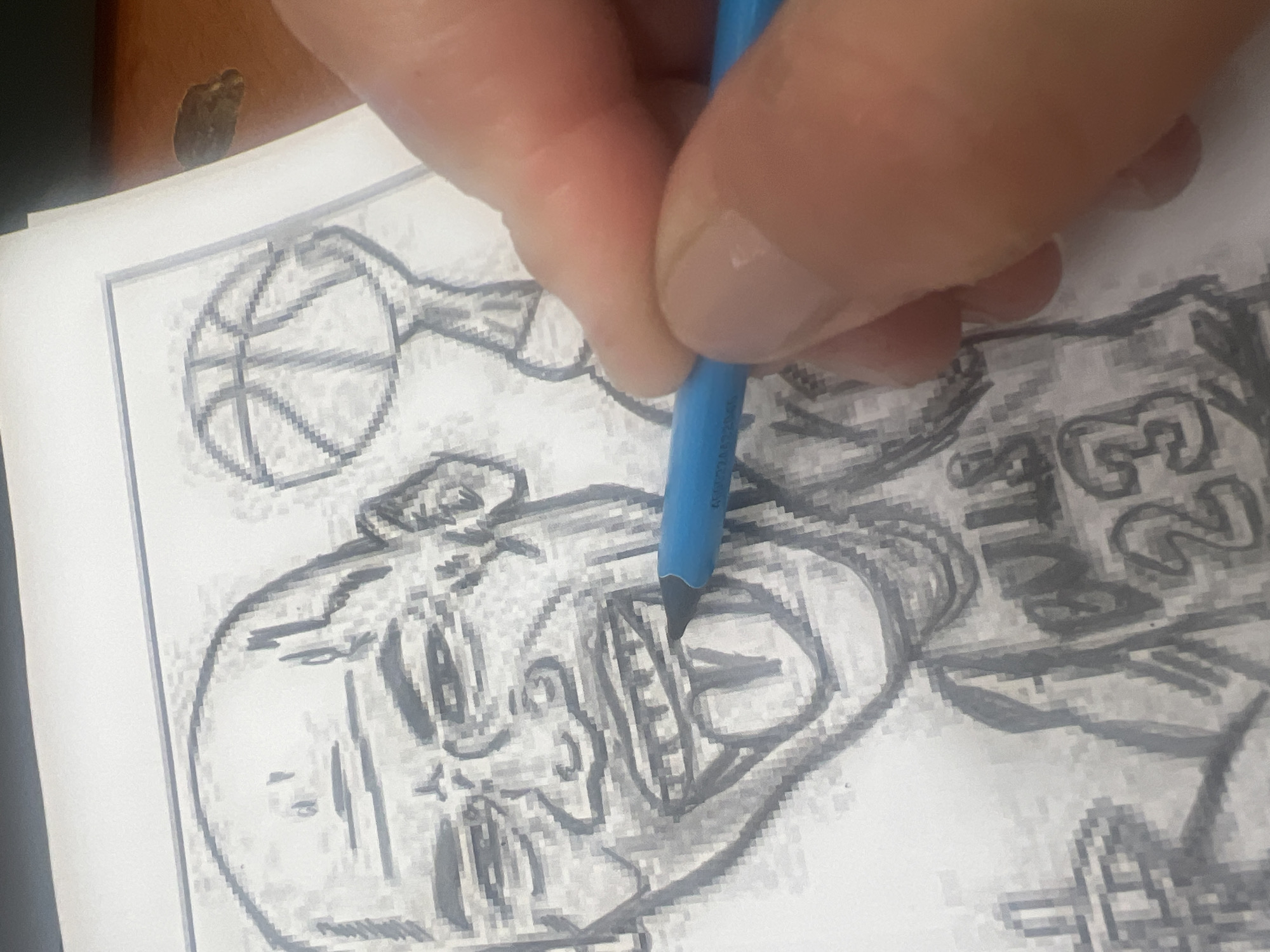 A hand holds a blue colored pencil, coloring in a drawing on white paper. The drawing appears to be of a basketball player, with a basketball visible. The number "23" is partially visible on the jersey. No information about Chaikovsky, Russia can be derived from this image.