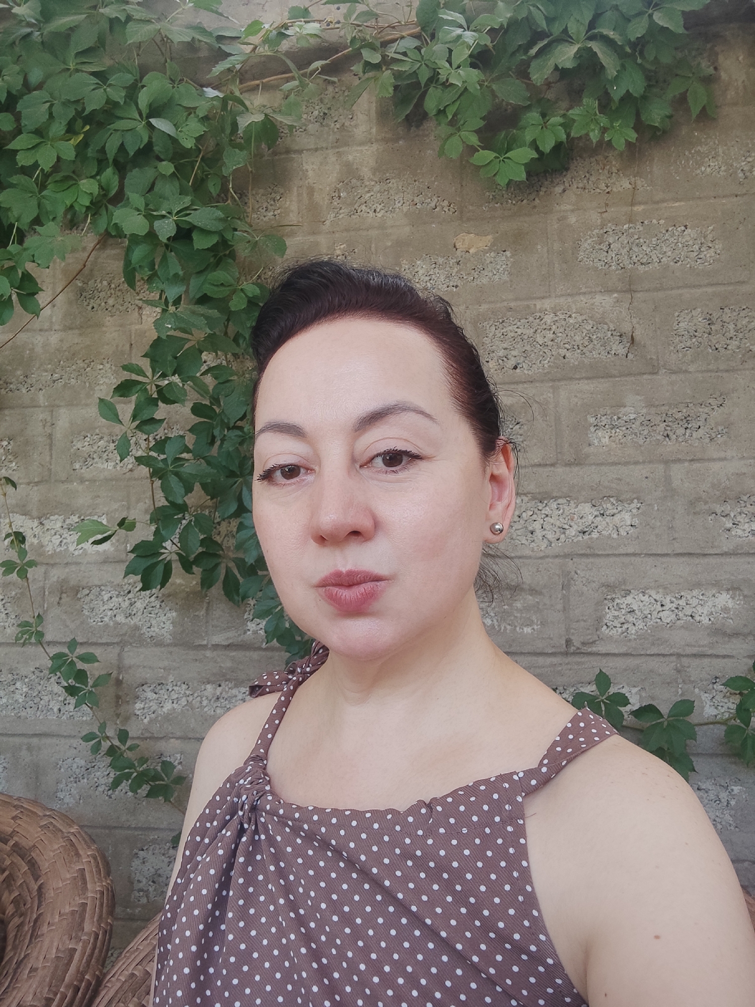 The image is a medium shot of a woman with dark hair pulled back, wearing a brown and white polka dot top with tie straps. She is looking directly at the camera with her lips slightly pursed. Her skin is fair, and she has light brown eyes. She is wearing a small silver stud earring in her right ear. The background consists of a textured, light-colored brick wall with green vines growing over it. To the left of the woman, partially visible, is a wicker chair. The lighting suggests it is daytime, possibly outdoors. The scene appears to be in a courtyard or garden area, potentially in Eshmataul, Uzbekistan.