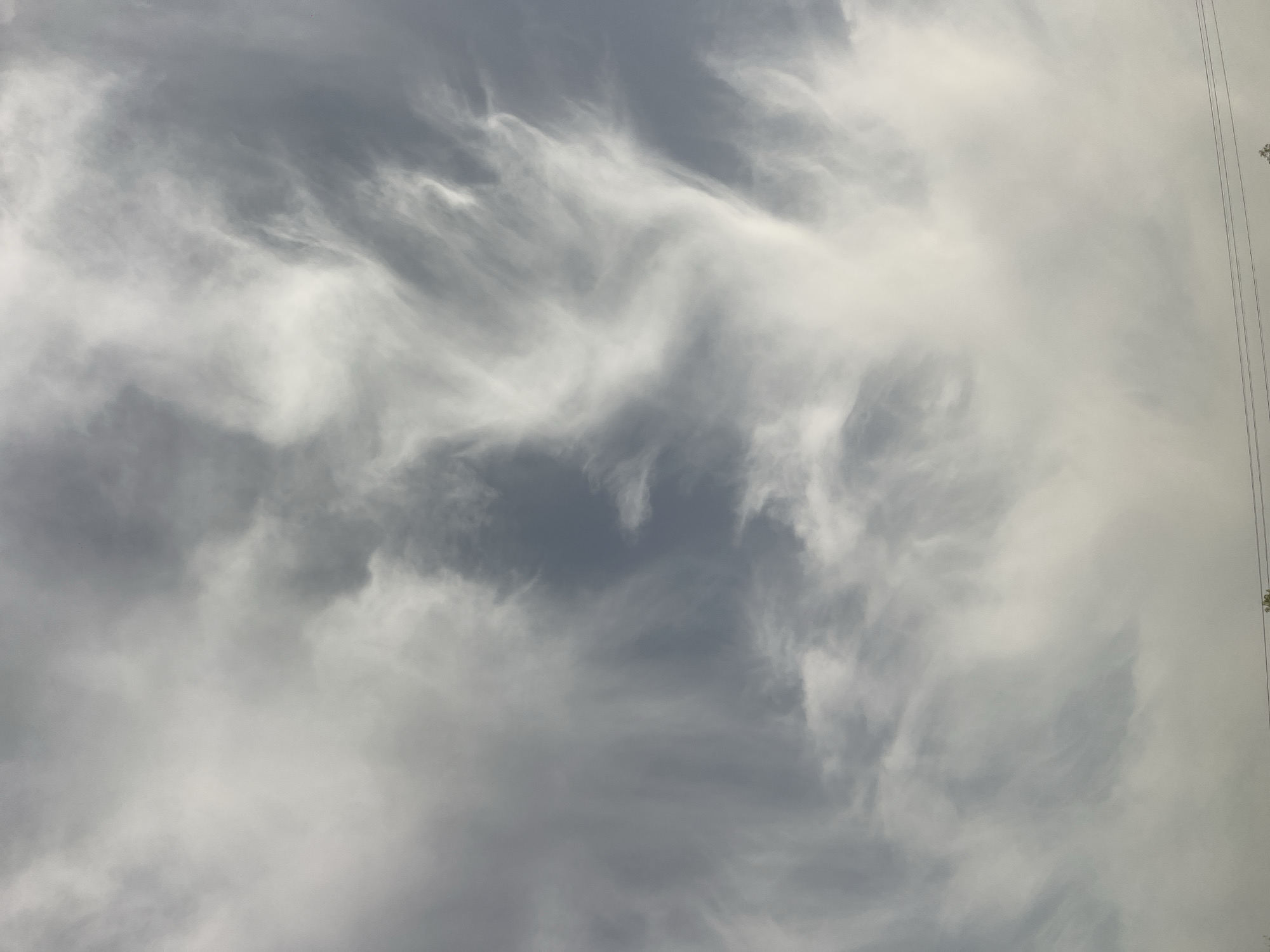 The image shows a cloudy sky in Howayi, Nigeria. The clouds are predominantly white and gray, with a wispy, textured appearance. There are no people visible. In the upper right corner, there are power lines and trees in the background.