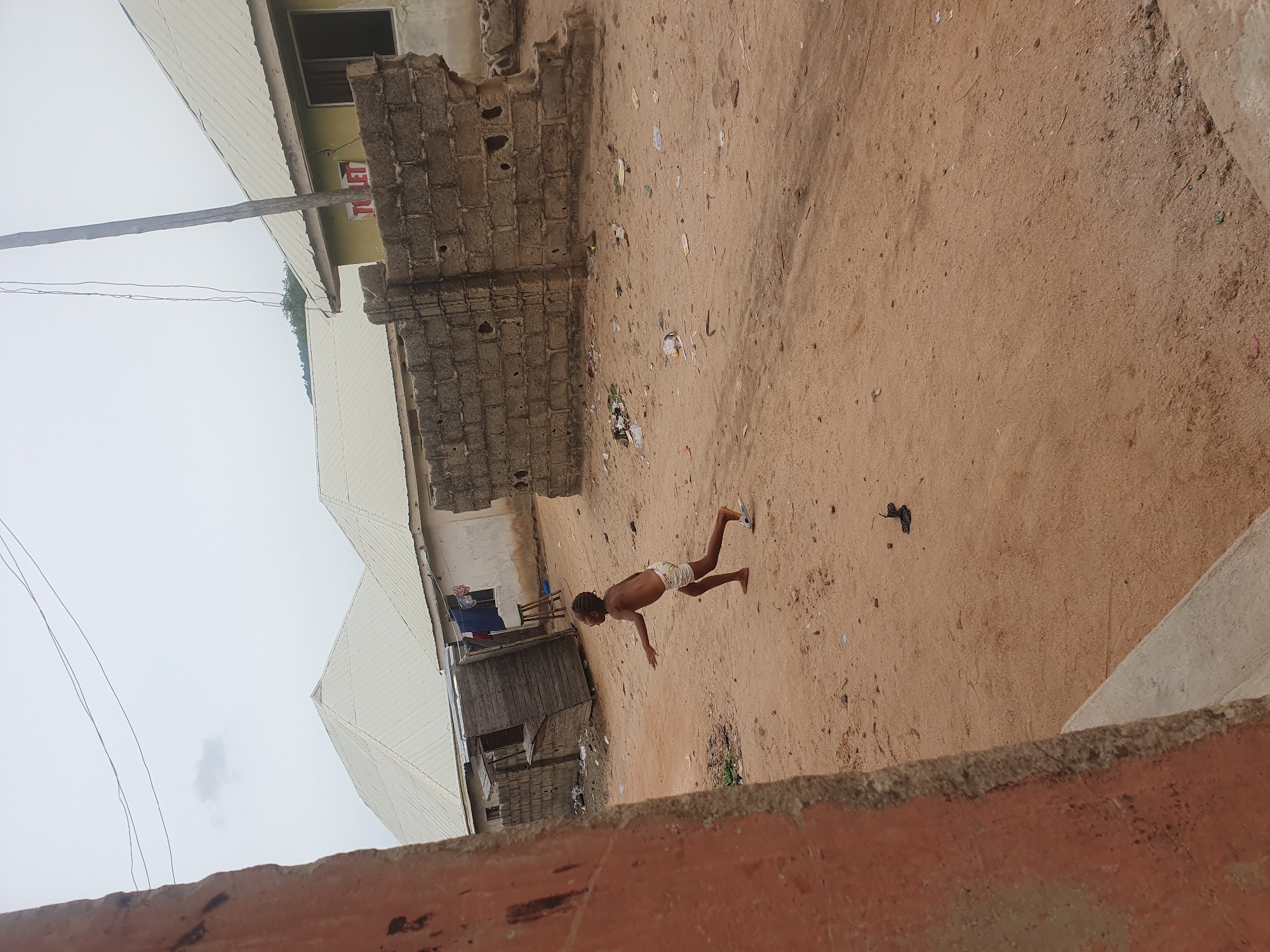 The image depicts an outdoor scene in what appears to be a low-income residential area in Lokoja, Nigeria. The environment is characterized by dirt ground and a mix of concrete and brick buildings. A young boy is playing in the dirt. Debris is scattered around. The architecture includes corrugated iron roofs, brick walls, and concrete structures. The image is taken from an elevated position looking downward. The sky is overcast.