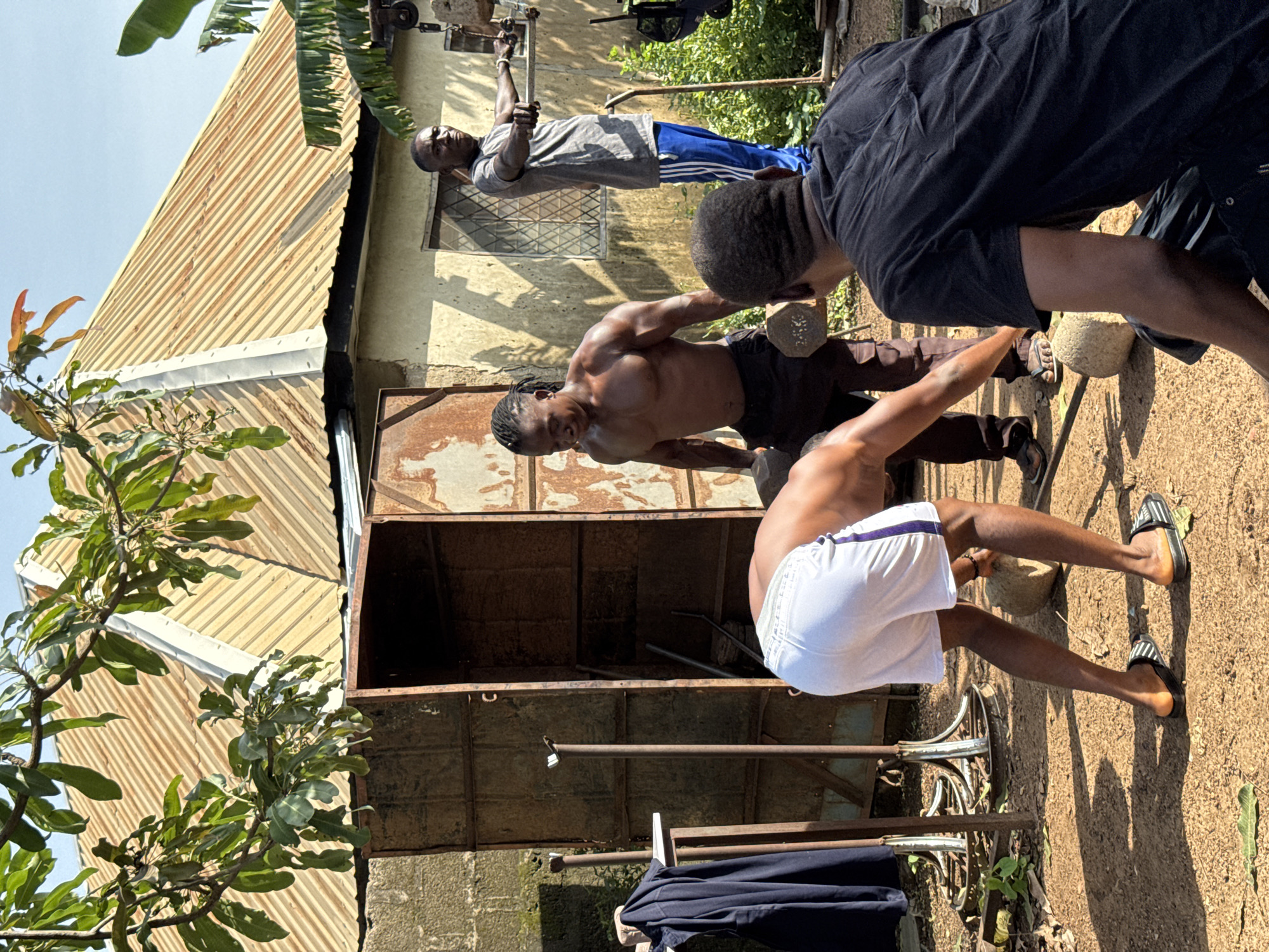 This media file depicts three individuals engaged in a workout session outdoors. The scene is set in Akwanga, Nigeria, with a building featuring a corrugated metal roof and a textured wall as the backdrop. Sunlight suggests it is daytime, and the overall ambiance points to warm weather.
The subjects are two men exercising and one man observing. One man, shirtless and wearing dark shorts, is performing a deadlift with a weight. Another man, also shirtless and wearing white shorts with purple trim, is in the process of performing a squat or lunge with a weight. The third individual, dressed in a grey t-shirt and blue athletic pants, is seen near some workout equipment in the background.
Notable details include the rustic workout equipment, likely made from repurposed materials, and the natural environment with foliage visible in the upper left corner. The ground is covered in dirt and gravel. The individuals appear focused on their exercise routine. No visible text is present in the media file.