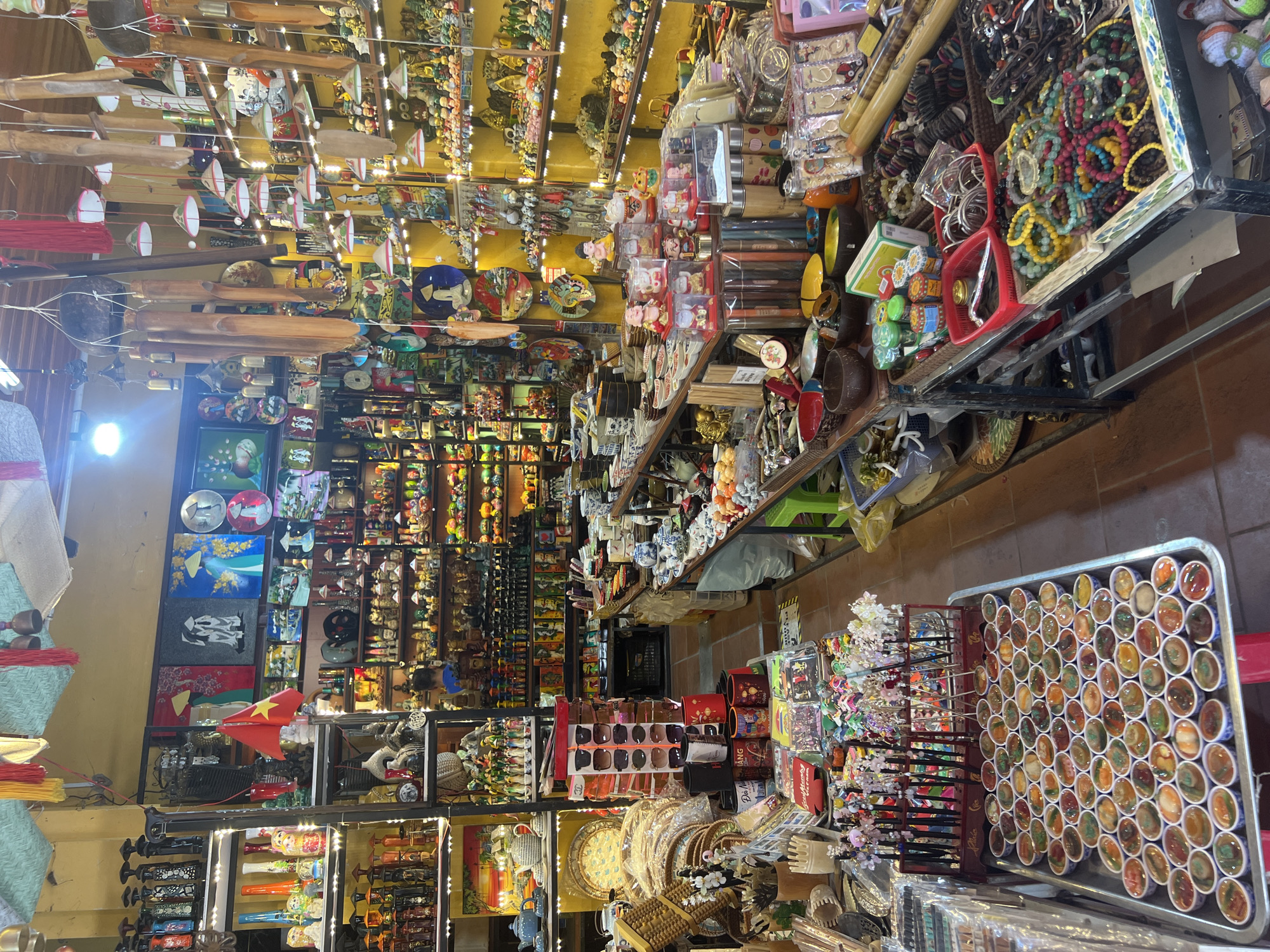 This image depicts a bustling souvenir shop in Hội An Ward, Vietnam. The shop is densely packed with a wide variety of merchandise, ranging from small decorative items to larger displays of ceramics, textiles, and wind chimes. The shelves are filled with numerous small figurines, including Buddhas and traditional Vietnamese characters, as well as intricately painted plates and bowls.
In the foreground, a large metal tray holds rows of small, colorful items that appear to be decorative stones or beads. To the right, a display rack showcases numerous bracelets made of various materials, including beads and what seem to be wooden or shell elements. Further back, tables are laden with more trinkets, including small potted plants, ceramic teacups, and packaged goods.
The shop's walls are painted a vibrant yellow, and a patterned ceiling hangs overhead, adorned with red tassels and wind chimes. A Vietnamese flag is prominently displayed on a shelf. While no explicit interactions are visible, the sheer volume of goods suggests a commercial environment catering to tourists. The lighting is a mix of natural and artificial sources, creating a warm and somewhat cluttered atmosphere. The floor is tiled with a reddish-brown hue.