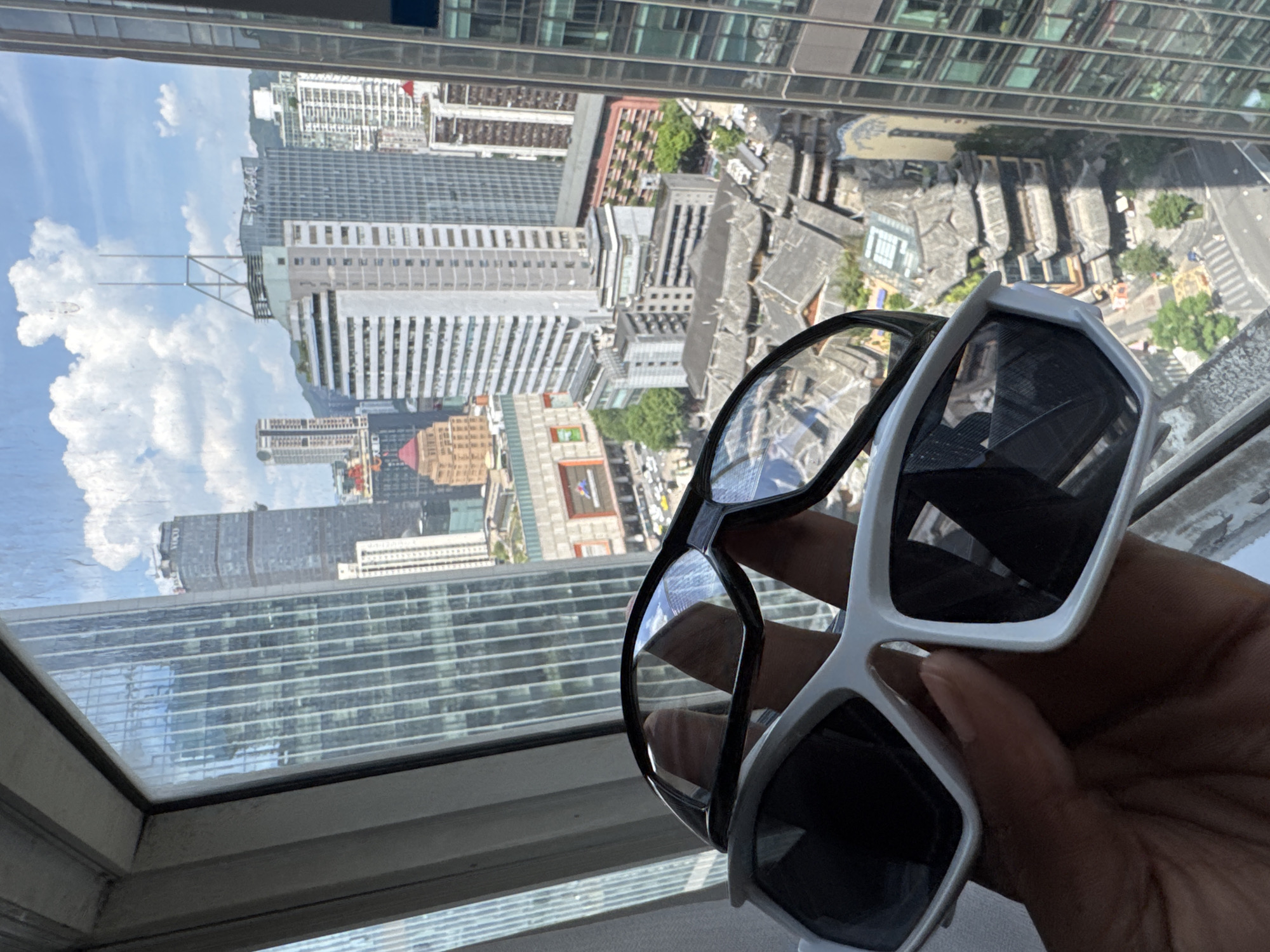 A person's hand holds up two pairs of sunglasses, a black-rimmed pair and a white-rimmed pair, in front of a window overlooking a cityscape in Yuzhong District, China. The sunglasses are positioned to partially obscure the view of the surrounding buildings. The cityscape features a mix of modern high-rise structures with glass facades and older, traditional Chinese architecture with tiled roofs. Lush green trees are interspersed among the buildings. The sky is blue with scattered white clouds. The image is taken from a high vantage point, suggesting the person is in a tall building.