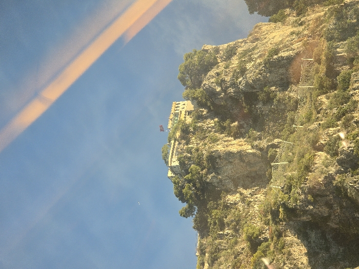 This image depicts a steep, rugged cliff face located in Beaulieu-sur-Mer, France, under a clear blue sky. A prominent light-colored, multi-story building with a flat roof and crenellated parapets is situated atop the cliff. A dark red flag with a lighter-colored emblem flies from a flagpole on the building's roof. The cliff face is characterized by exposed rock formations and dense green vegetation, including numerous trees and shrubs clinging to the slopes. Multiple sections of light-colored rockfall netting, supported by poles, are visible across the mid and lower portions of the cliff, indicating slope stabilization or erosion control measures. Direct sunlight illuminates the scene from the left, casting highlights and shadows across the rocky terrain. The upper right quadrant of the image displays two distinct, parallel, diagonal orange-yellow streaks, consistent with reflections on a transparent surface. Several smaller, amorphous white light reflections are present in the lower left quadrant. No individuals or active subjects are present in the scene.