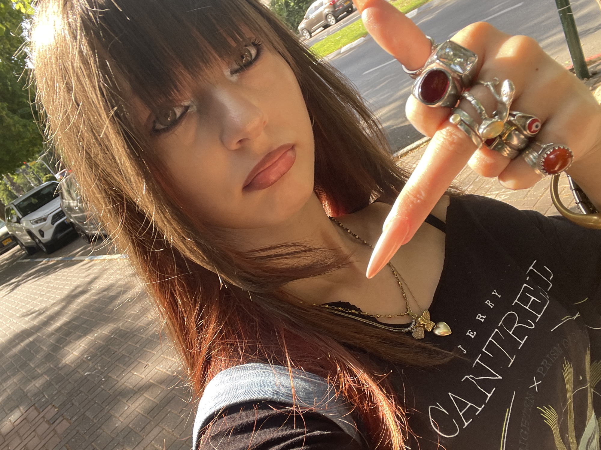 A young woman with long, reddish-brown hair and bangs is taking a selfie outdoors, with a street and sidewalk visible in the background. She is wearing a black tank top with the text "JERRY CANTREL" printed on it and a denim garment. She is adorned with multiple rings on her fingers, including one with a large red stone and another with an evil eye design. She also wears several necklaces, one with a golden heart pendant. The lighting suggests it is daytime, with strong sunlight creating shadows. The street appears to be in Raanana, Israel, based on the presence of cars and road markings typical of the city.