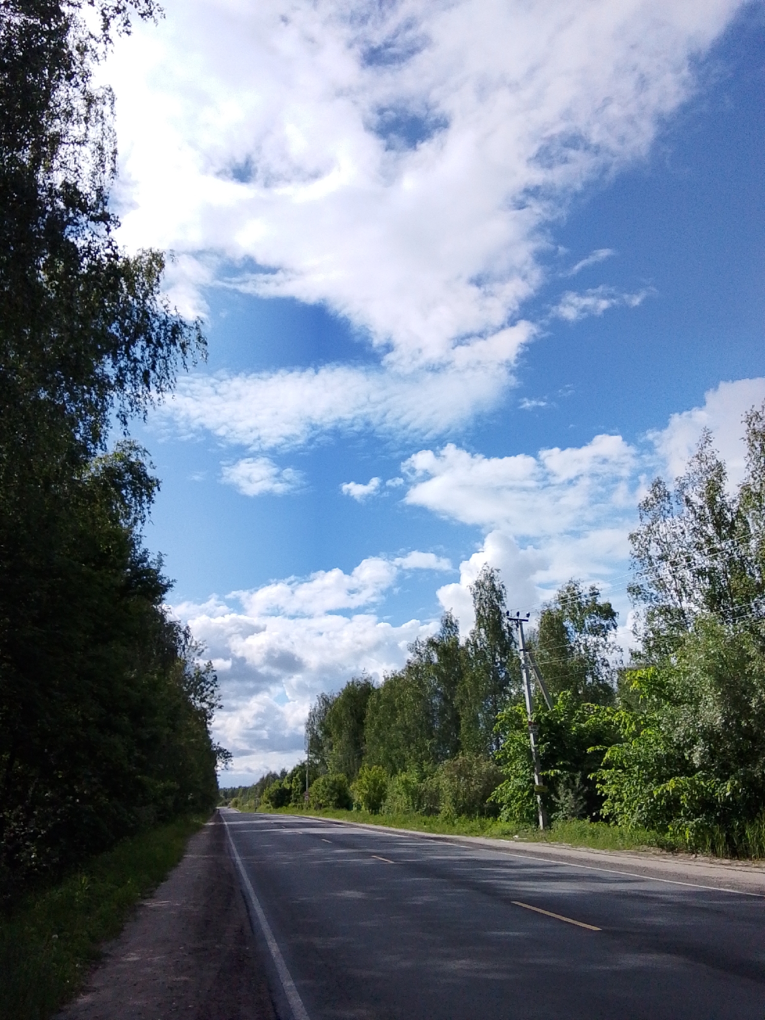The image shows an asphalt road in Снопок Новый, Russia. The road stretches into the distance between lines of trees on both sides. The sky is bright blue with scattered white clouds. There are power lines and poles visible along the right side of the road. The road has a yellow line marking the lanes. There is a narrow dirt and gravel shoulder on the left side of the road. No people or animals are visible.