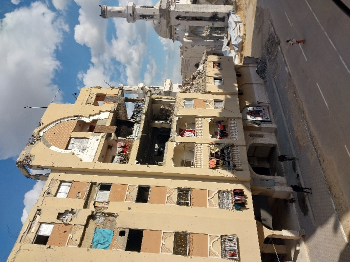 The image depicts a street scene in Khan Yunis, Palestinian Territory, under a partly cloudy sky. The prominent structure on the left is a severely damaged multi-story building, approximately five to six floors high, with a tan/beige facade. Its upper floors and central section exhibit extensive structural collapse, revealing exposed interiors, twisted rebar, and rubble. A large, decorative arched segment on the upper facade is heavily damaged. Numerous circular impact marks are visible across the building's exterior. Clothes are hanging from windows and balconies on the lower floors, some of which appear relatively less damaged.
To the right, partially obscured by the foreground building, stands the Al-Farouq Mosque, identified by its white dome and tall, slender minaret. Both the mosque's main structure and its minaret show significant damage, with visible erosion and destruction to architectural elements.
In the foreground, an asphalt road with a white centerline extends across the scene. Adjacent to the damaged building is a paved sidewalk. Three individuals are visible on the street: two men in dark clothing walk on the sidewalk near the damaged building, and one person, dressed in orange pants and a white top, walks in the middle of the road further to the right. Piles of debris and rubble are present on the sidewalk and in an open dirt area between the damaged building and the mosque. Several makeshift shelters, constructed with tarpaulins, are visible in the cleared ground near the mosque. The sky is blue with scattered white cumulus clouds, indicating daytime lighting.