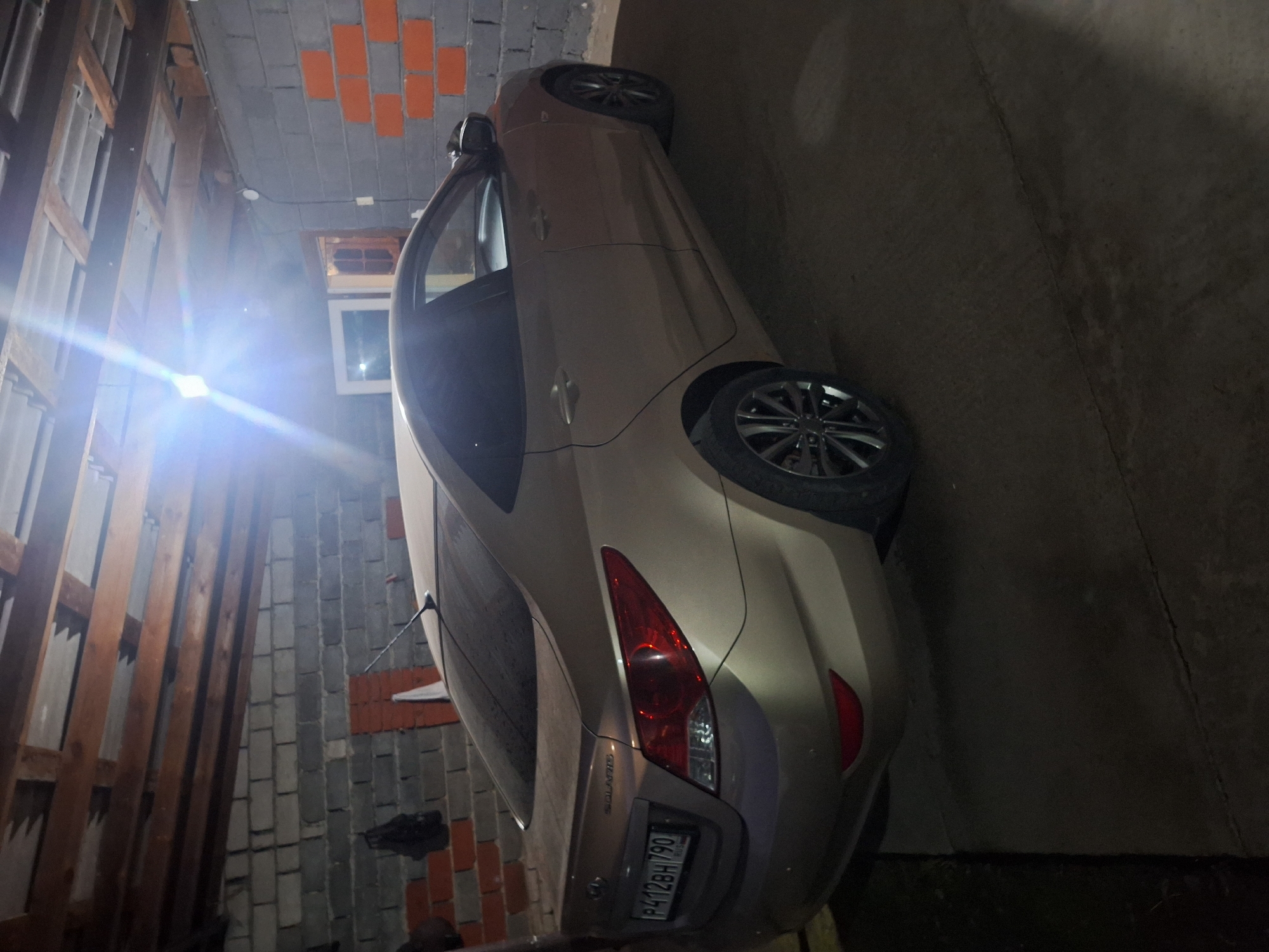 A gold Hyundai Solaris sedan is parked in a garage. The car is viewed from the rear-left quarter. The license plate reads "P412BH 790". The garage has concrete floors and walls made of cinder blocks and some orange bricks. Wooden beams are visible on the ceiling. A bright light source is visible in the upper portion of the image, causing glare. There is no visible indication of the city of Chermen, Russia.