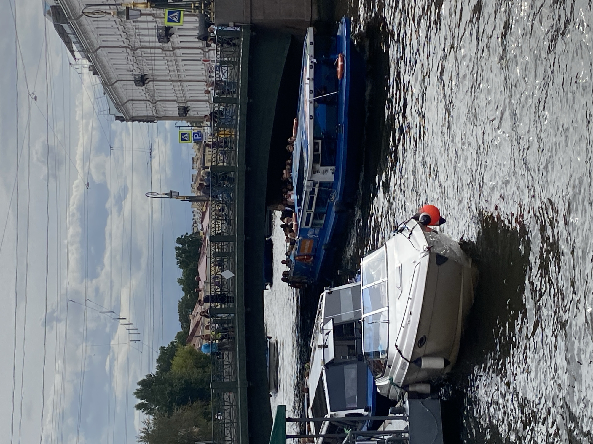This image depicts a canal in Saint Petersburg, Russia, likely the Griboyedov Canal, given the iconic architecture and bridge visible. Two tour boats are navigating the waterway under a green bridge. The larger, blue boat on the right is filled with passengers, suggesting a guided tour. A smaller, white motorboat is docked on the left, also appearing to be engaged in tourism. People are visible on the bridge, observing the boats and the surroundings. The buildings lining the canal showcase the classical architectural style characteristic of Saint Petersburg. Overhead, a complex network of power lines crisscrosses the sky, a common sight in urban environments. The water is rippled, indicating movement and the presence of other vessels. The sky is partly cloudy, with a moderate amount of daylight filtering through.