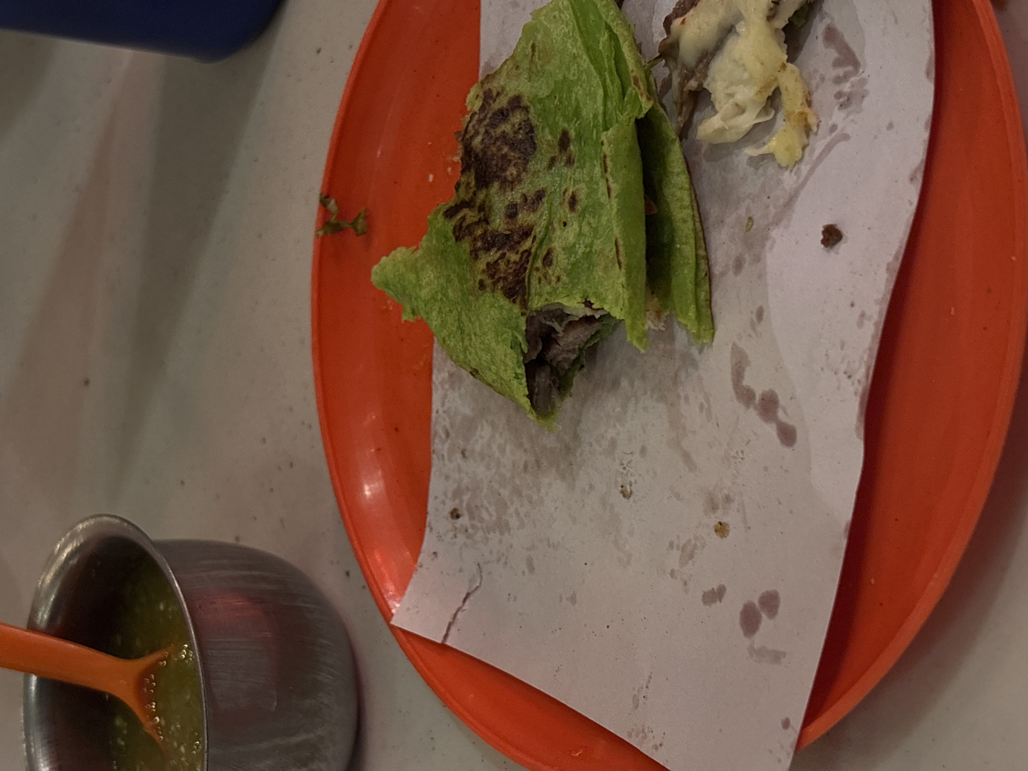 The image shows a partially eaten taco on an orange plate, placed on what appears to be a table. The taco is wrapped in a green tortilla and contains meat. There is a piece of white paper beneath the taco. A small metal bowl containing green salsa and an orange spoon are visible to the left of the plate. The setting appears to be a restaurant. While not verifiable with certainty, based on the food style (taco), the image is potentially taken in Mexico City, Mexico.