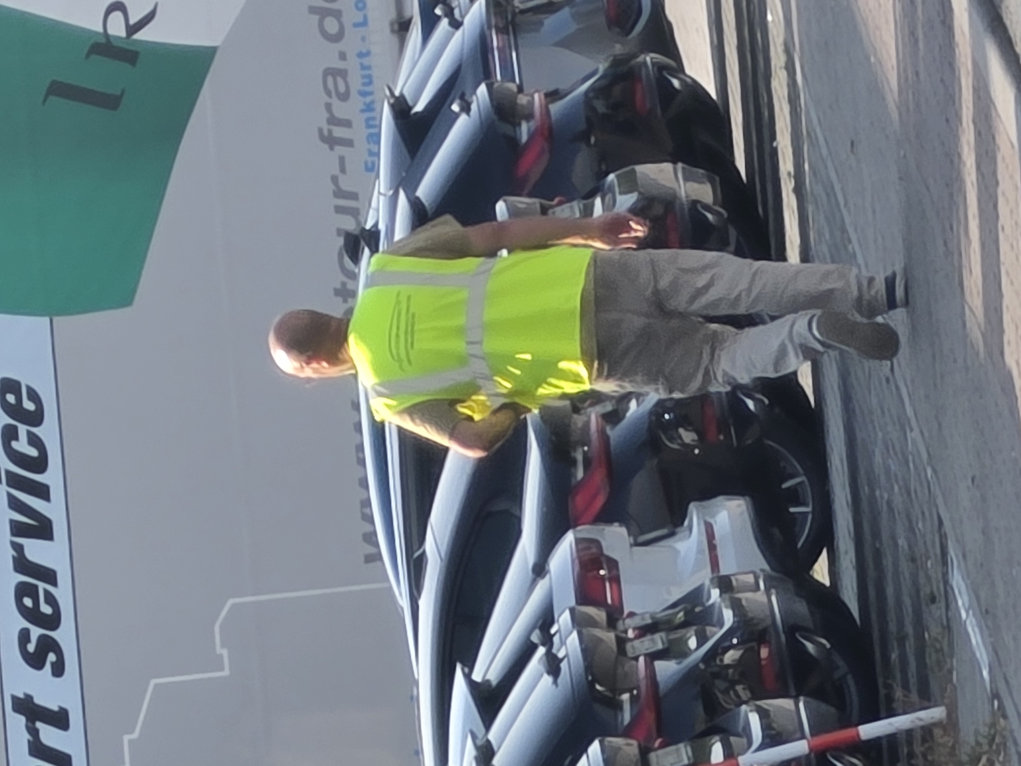 A bald man wearing a bright yellow safety vest and light-colored pants walks away from the camera. He is in a lot with many cars. The man is walking on a paved surface. In the background, there is a large white truck with green lettering. The lettering appears to say "tour-fra.de" and "Frankfurt - Lo". The cars are dark-colored sedans parked in rows. The scene is illuminated by sunlight, casting shadows on the ground.