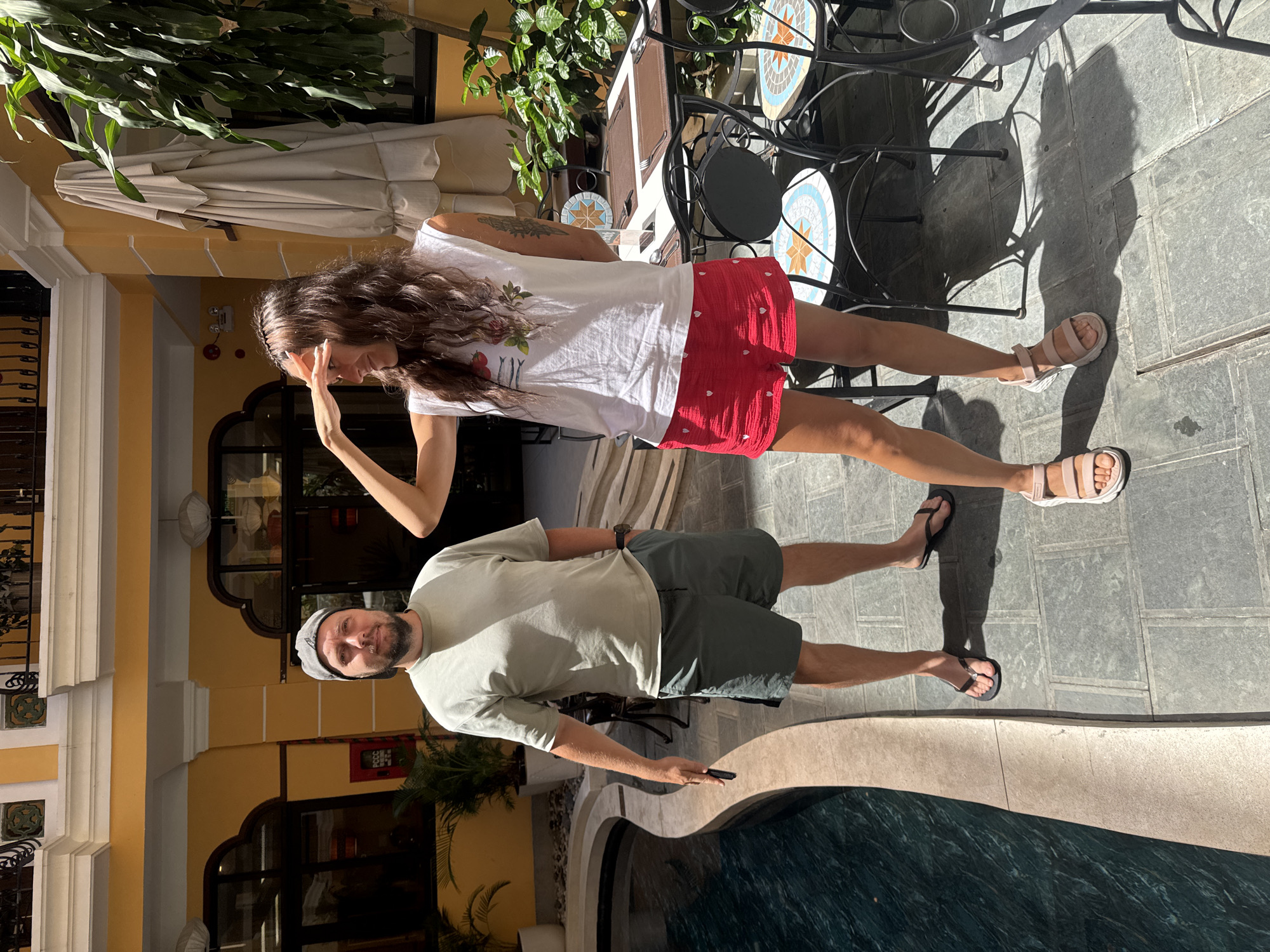 A man and a woman stand on a tiled patio next to a pool. The woman wears a white t-shirt, red shorts, and sandals, and she has her hand raised to her forehead. The man wears a gray hat, a light green t-shirt, dark green shorts, and flip-flops. The patio has tables with black metal frames and decorative tile tops. The building in the background is painted yellow and has arched windows. It may be in Đà Nẵng, Vietnam.
