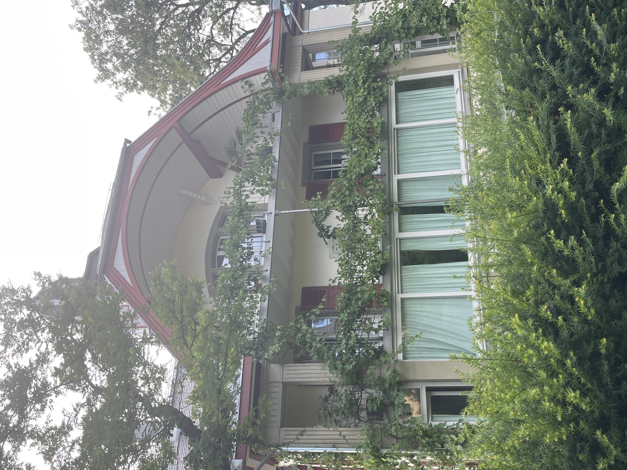 The image shows a multi-story building with a red-trimmed arched roof, situated in Bern, Switzerland. The building features several windows, balconies, and is partially obscured by lush green trees and bushes. One balcony on the second floor is adorned with climbing vines and has a railing. Red shutters are visible on some of the windows. Light green curtains cover the windows on the first floor. The overall setting suggests a residential or possibly a historical building. The sky is overcast, contributing to a soft, diffused light.