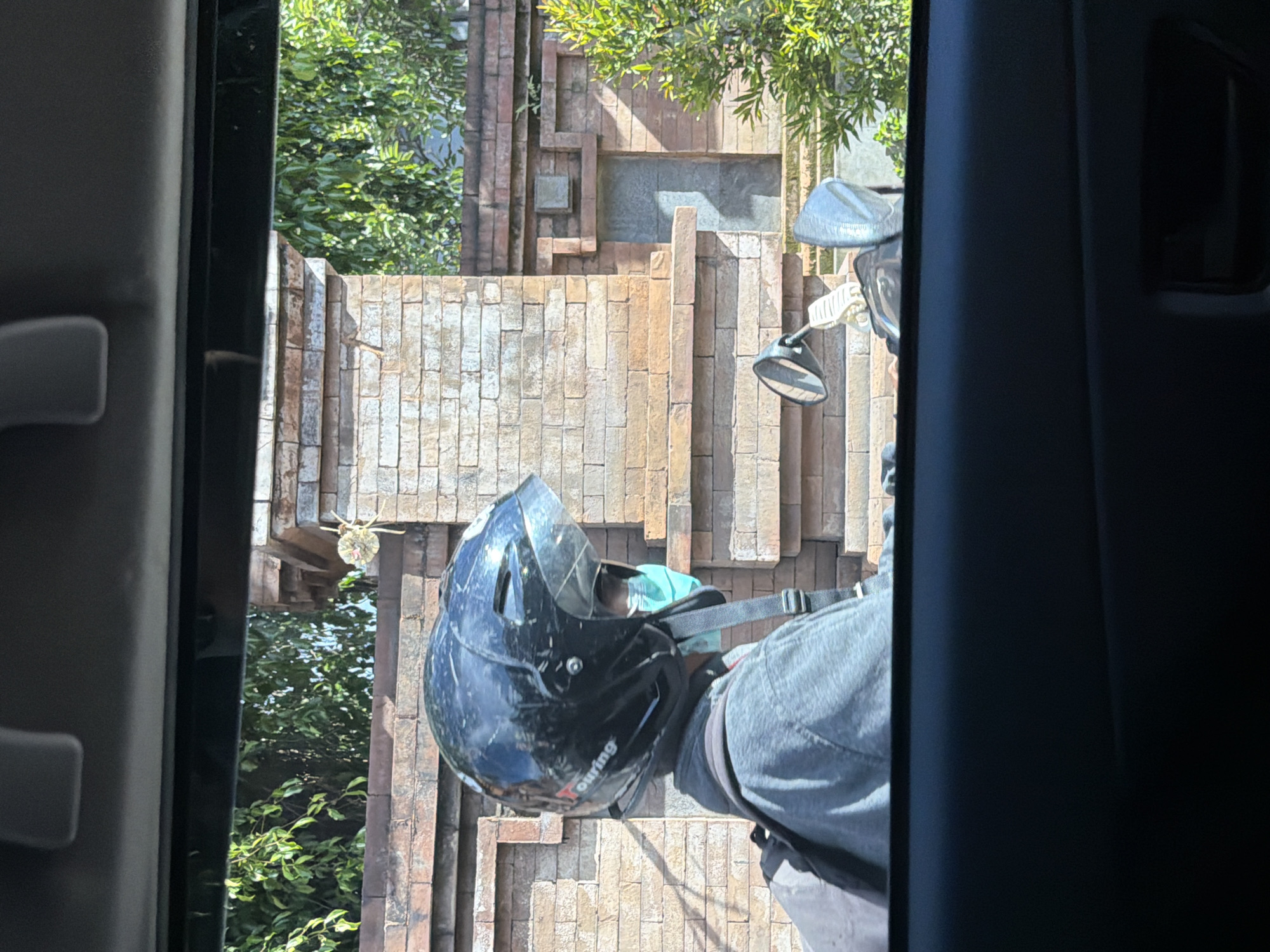 The image, captured from within a vehicle, looks out through an open door or window, framing a motorcyclist and a detailed brick wall under bright daylight. A person is seen from the chest up, wearing a black full-face motorcycle helmet with a clear visor, which shows signs of wear and scratches. A light blue surgical mask is visible beneath the helmet's chin strap. The word "Touring" is faintly discernible in white text on the side of the helmet. The person's left arm, clad in a grey long-sleeved garment, is partially visible, along with what appears to be a dark pant leg or backpack strap. To the right of the person, parts of a motorcycle are visible, including a silver fairing or handlebar cover and a black rearview mirror.
The background features an intricately designed wall constructed from various shades of brown and beige bricks or stone blocks. The wall exhibits a multi-dimensional pattern with recessed and protruding sections, including a rectangular inset in the upper central area. Lush green foliage from trees and bushes is visible above and behind the wall, suggesting an outdoor, tropical setting consistent with North Kuta, Indonesia. The bright sunlight casts distinct shadows, indicating clear weather during the day. The motorcyclist appears to be stationary, possibly waiting.