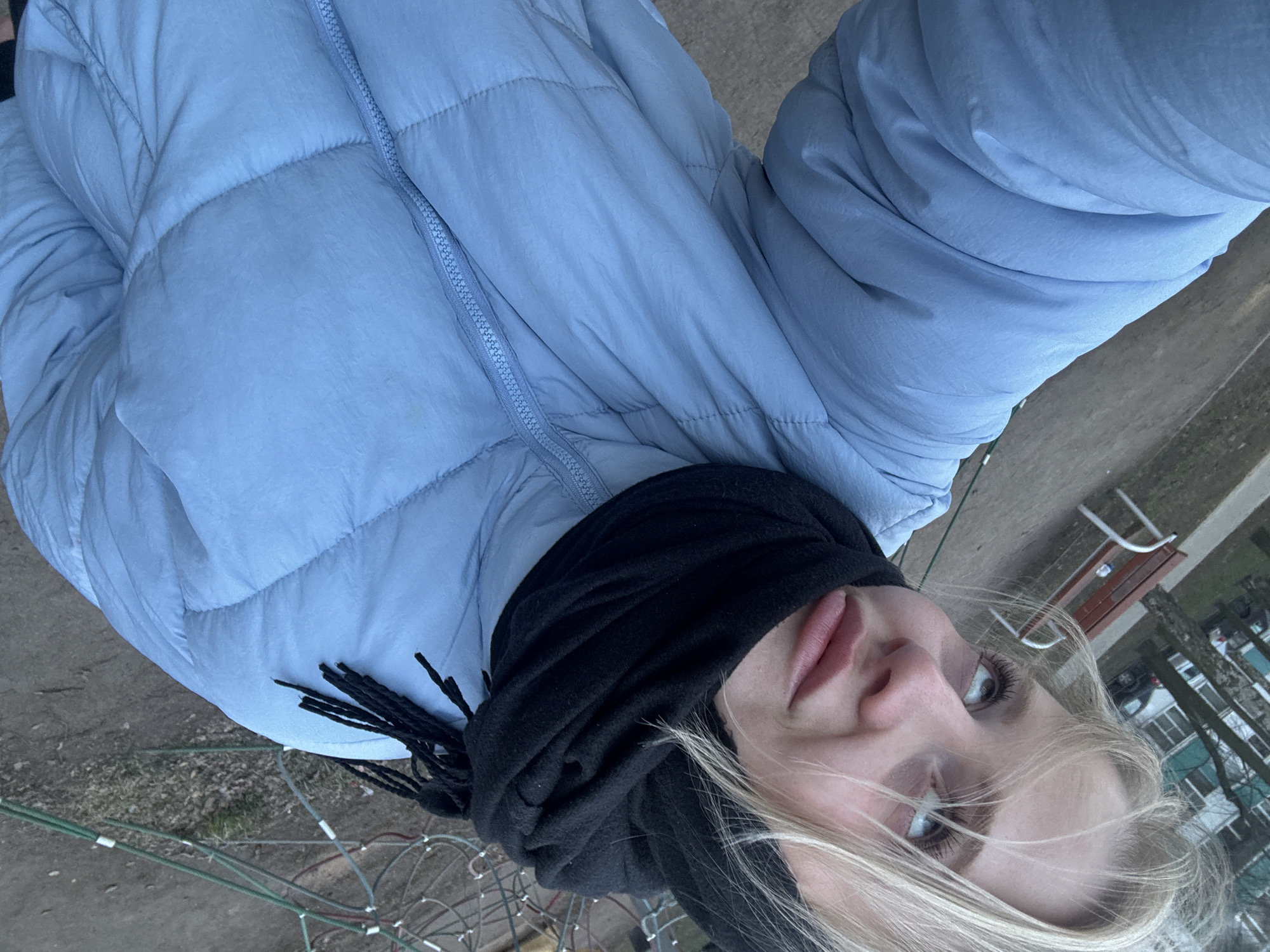 A selfie was taken outside in Saint Petersburg, Russia. A young woman with light blonde hair and fair skin is wearing a light blue puffer jacket and a black scarf. The background shows a park with benches, trees, and what appears to be playground equipment with intertwined ropes. The ground is a mix of dirt and grass.