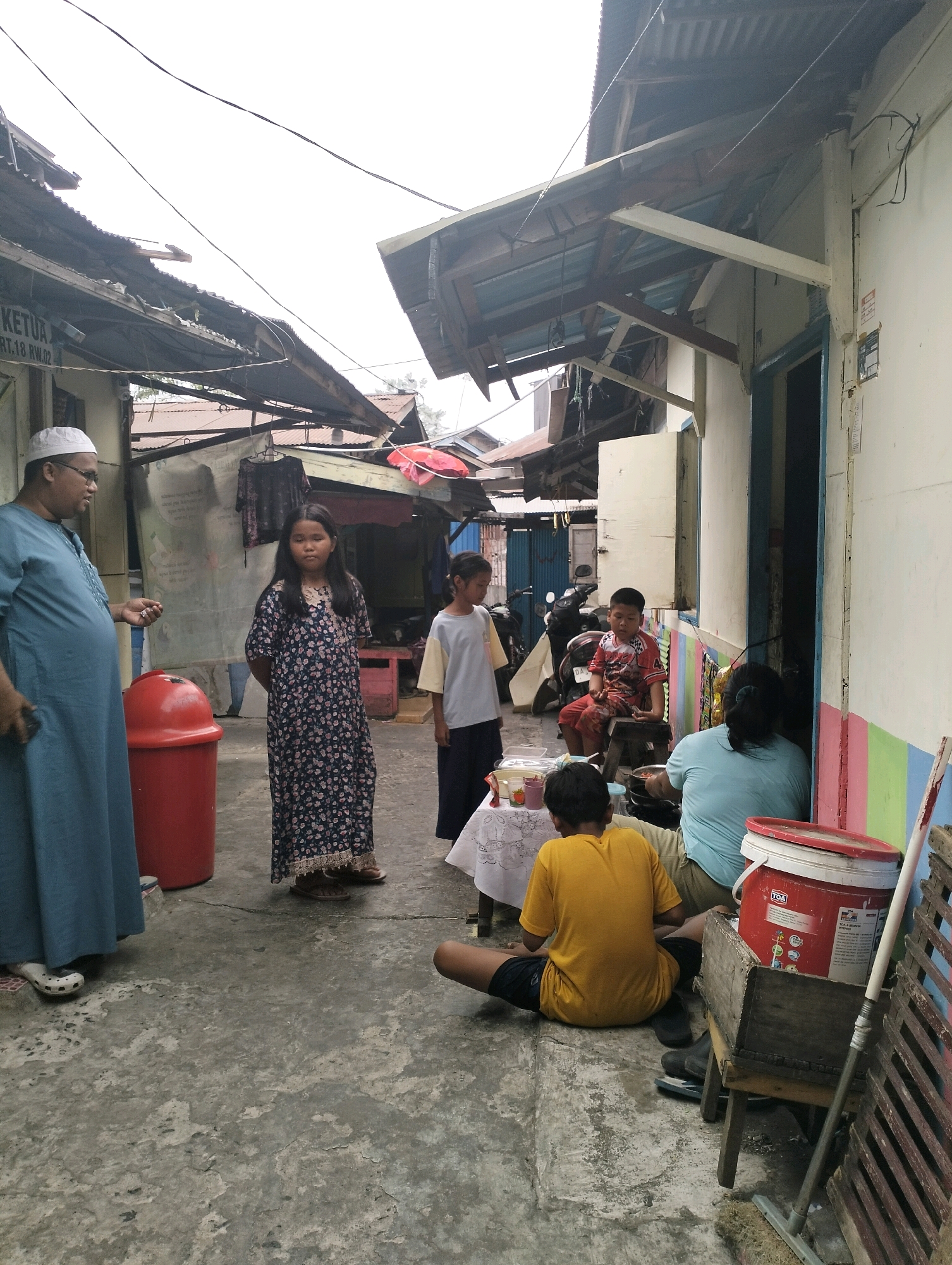 The image depicts a street scene in Banjarmasin, Indonesia. In the foreground, a man wearing a blue robe and a white cap, possibly a community leader, is engaged in a conversation with a young girl in a floral dress. Beside them is a large red trash can. Further back, other individuals are gathered. A boy in a red and white shirt is seated on a stool, while a girl in a light-colored t-shirt and dark pants stands near a table with food containers. To the right, two women are sitting, one of whom is wearing a light blue shirt. The environment appears to be an alleyway with a row of buildings on the right, featuring a blue door and colorful stripes on the wall. Overhead, exposed electrical wires crisscross the sky, and the roofs of some structures show signs of disrepair. In the background, motorcycles are parked. The ground is paved with concrete, showing signs of wear. A red bucket with writing on it and a broom are visible on the right side of the frame.