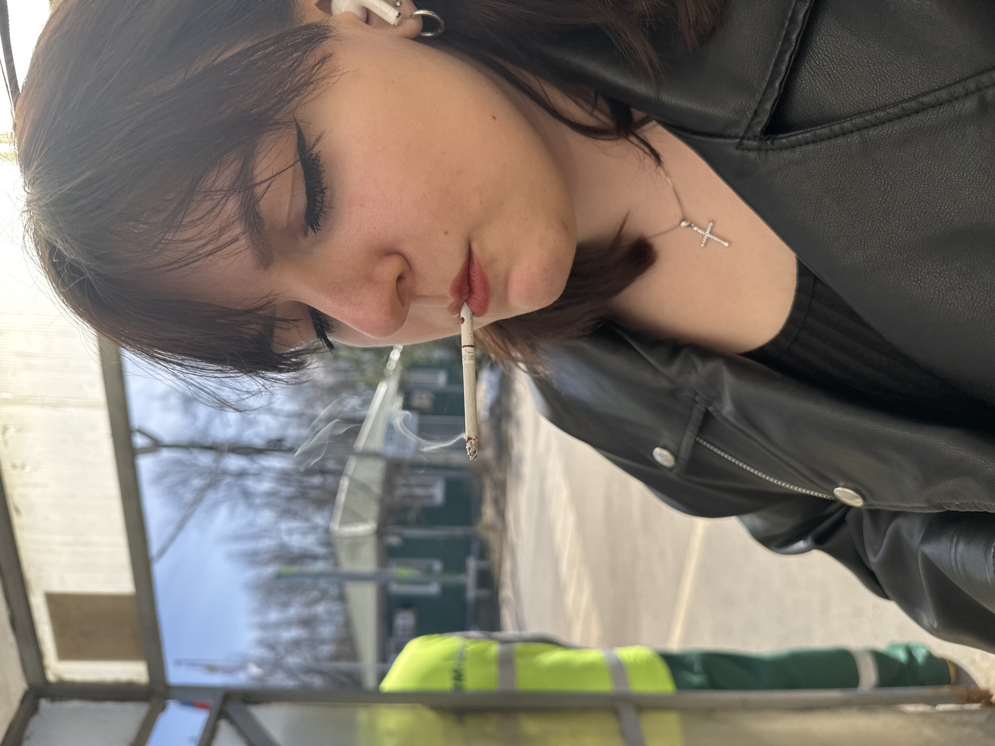 A young woman with dark hair, wearing a black leather jacket and a silver cross necklace, holds a lit cigarette in her mouth. She has closed eyes and is positioned at an angle to the camera. She is wearing earbuds in her left ear. Behind her is a blurred background of trees and a building with green accents. The scene appears to be in an outdoor setting, possibly a bus stop, with a yellow and green object visible in the foreground. The photograph was taken in Dmitrov, Russia.
