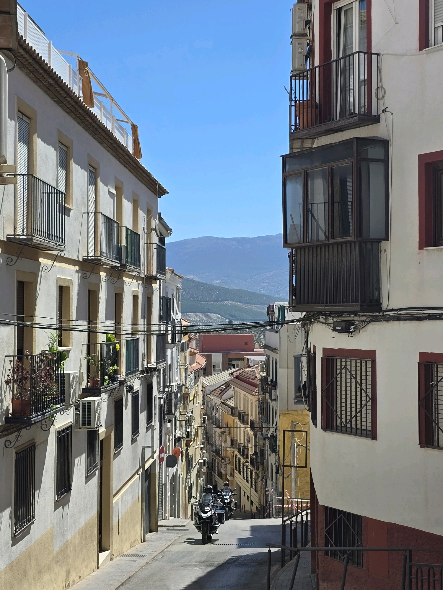 The image depicts a steep, narrow street in the historic quarter of Jaén, Spain, lined with traditional whitewashed buildings adorned with wrought-iron balconies and terracotta roof tiles. The architecture suggests a Mediterranean style, common in Andalusian cities. The buildings on either side of the street feature multiple stories, with windows protected by decorative grilles. Air conditioning units are visible mounted on the exterior walls of some apartments.
The street itself is paved with cobblestones and slopes downwards, creating a sense of depth and perspective. Several motorcycles, with riders, are navigating the incline, suggesting active local transportation within the old city. In the background, a hazy mountain range, possibly the Sierra de Jaén, forms a backdrop against a clear blue sky. Olive groves or vineyards are visible on the lower slopes of the mountains, indicating the agricultural significance of the region. The sunlight casts strong shadows, highlighting the textures of the buildings and the uneven terrain. The overall atmosphere is one of a sun-drenched, historic urban environment.