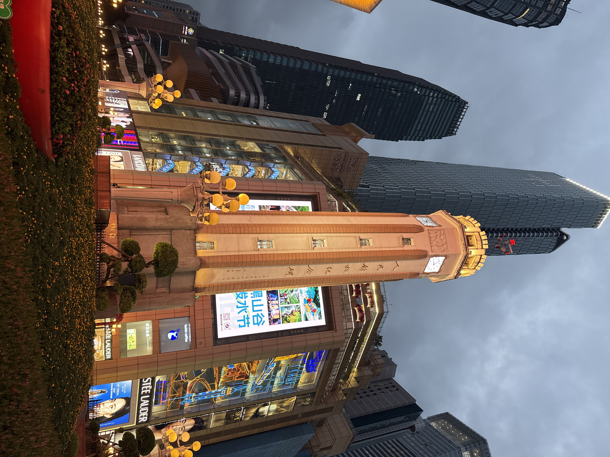 In the Yuzhong District of China, this image captures a cityscape dominated by modern skyscrapers against a cloudy twilight sky. A prominent, illuminated clock tower stands as a focal point, adorned with ornate carvings and vertical text. The tower is attached to a multi-story building featuring large digital billboards displaying advertisements, some with vibrant neon accents. Storefronts such as "STEE LAUDER" and "LANCOME" are visible, indicating a commercial district. The foreground is filled with a meticulously landscaped garden featuring low-lying flowering plants and decorative trees, enclosed by a black metal fence. Streetlights and decorative lamps with multiple bulbs illuminate the scene, casting a warm glow on the surroundings. The overall impression is one of a bustling, modern urban environment.