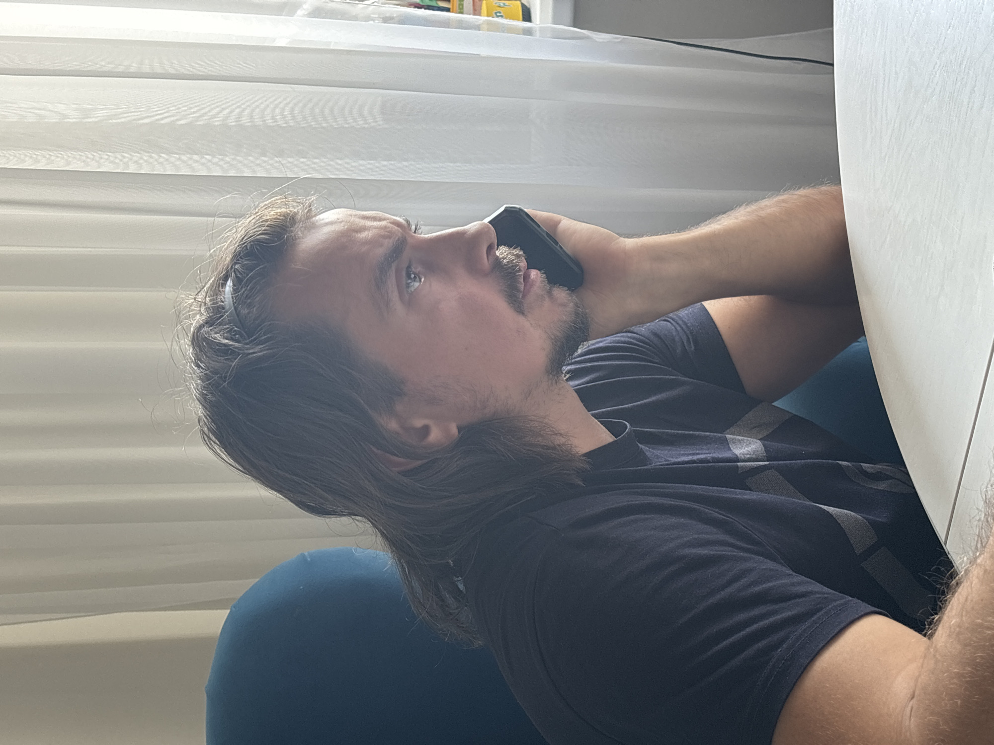 A man with long brown hair and a goatee is talking on a black smartphone. He is wearing a dark blue t-shirt and sitting on a blue, round chair. He is positioned next to a white table or desk. The background features a white, horizontally-lined window covering. Location: Unknown, Unknown.
