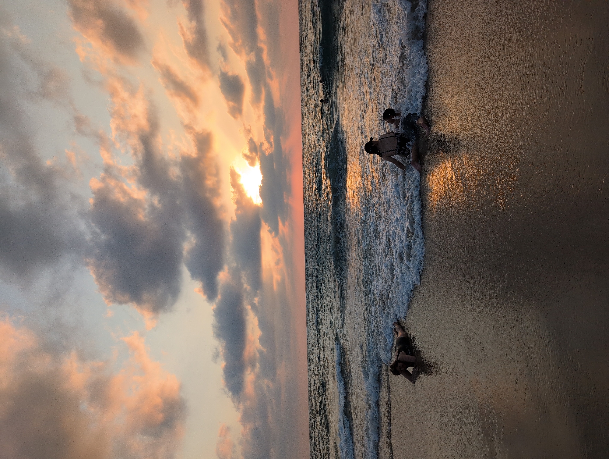 The image captures a sunset scene at Palmachim Beach, Israel. Three individuals are visible on the wet sand near the waterline. On the left, a person is lying prone facing the sand with their head turned towards the viewer. In the center, two individuals are seated with their legs extended into the shallow water, being splashed by waves. The water is dark and choppy, with white foam from the breaking waves. In the distance, several other figures can be seen in the ocean. The sky is filled with dramatic clouds, tinged with orange and yellow from the setting sun, which is partially obscured by clouds. The wet sand reflects the colors of the sky and the ocean.