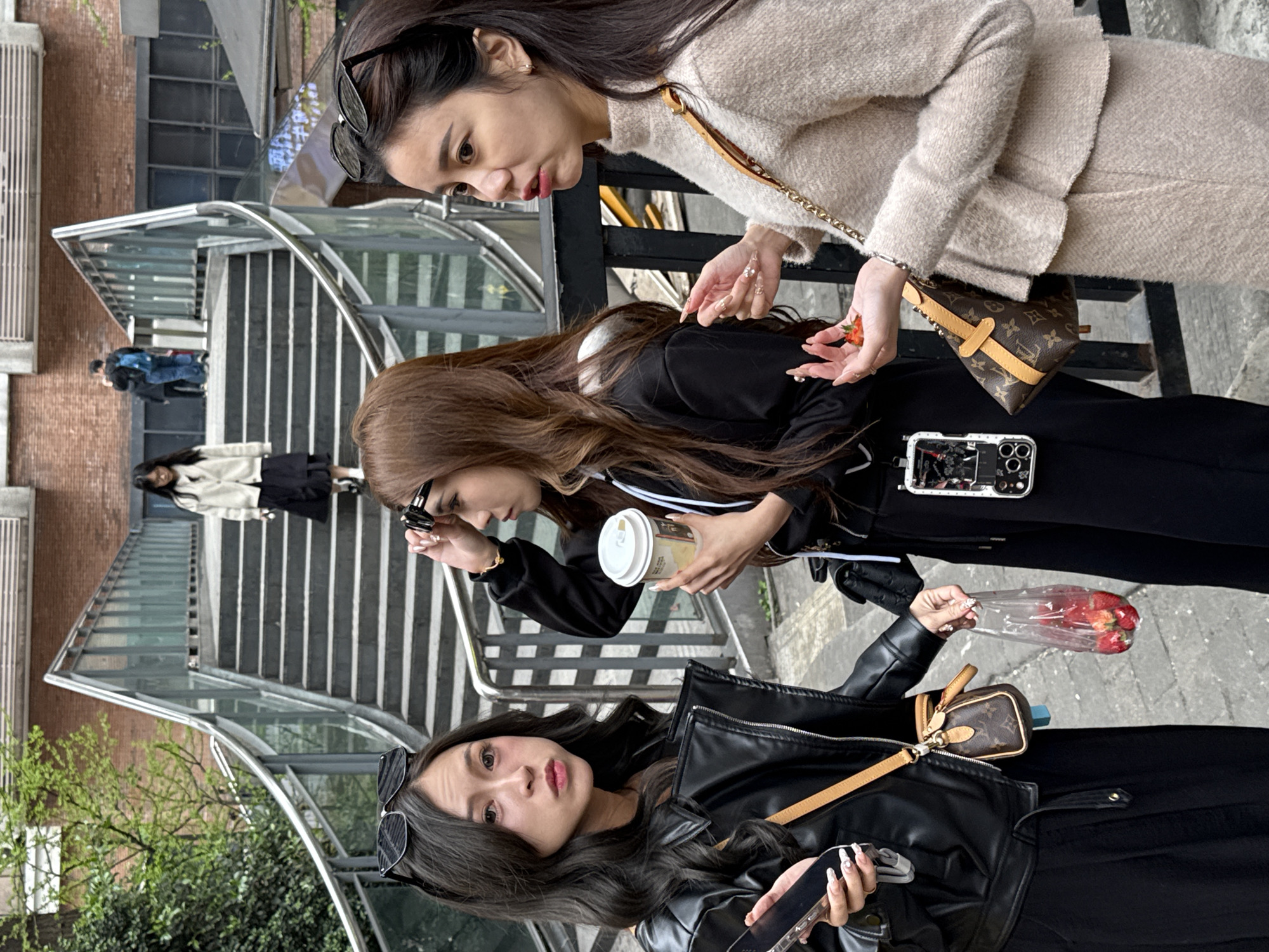 This image depicts an outdoor urban scene in Yuzhong District, China, featuring five individuals.
In the foreground, three young adult East Asian females are positioned near a black metal barrier.
1. On the left, a female with long dark hair, wearing a black faux leather jacket over dark clothing, has black sunglasses pushed onto her head. She holds a dark smartphone in her right hand and a clear plastic bag containing approximately six red strawberries in her left hand. A small brown Louis Vuitton monogrammed bag with a tan strap hangs from her right shoulder. Her lips are slightly pursed as she looks forward.
2. In the center, a female with long brown hair, wearing a black hooded top with white trim and black trousers, holds a white disposable coffee cup with a lid in her left hand. Her right hand adjusts a pair of dark rectangular sunglasses on her forehead. A white smartphone in a clear decorative case is clipped to her left hip. She is looking towards the female on her right. Her left hand is in contact with the plastic bag of strawberries held by the first female.
3. On the right, a female with long dark hair, wearing a light beige textured knitted sweater and matching skirt, holds approximately three red strawberries in her cupped left hand. Her right hand is slightly raised with an open palm. A small brown Louis Vuitton monogrammed bag with a gold chain strap is slung over her left shoulder. Her mouth is partially open as she looks towards the center female.
In the background, a concrete staircase with a curved stainless steel handrail and glass panels ascends and descends. A female with dark hair, wearing a light-colored cardigan and dark skirt, is visible descending the stairs. Further up the stairs, two male figures are seen ascending; one wears a dark jacket and a blue backpack, while the second is partially obscured behind him.
The environment includes green foliage on the far left, a red brick building with ventilation grates above the stairs, and a dark structure with windows beneath the brick building. The immediate foreground consists of a grey paved surface.