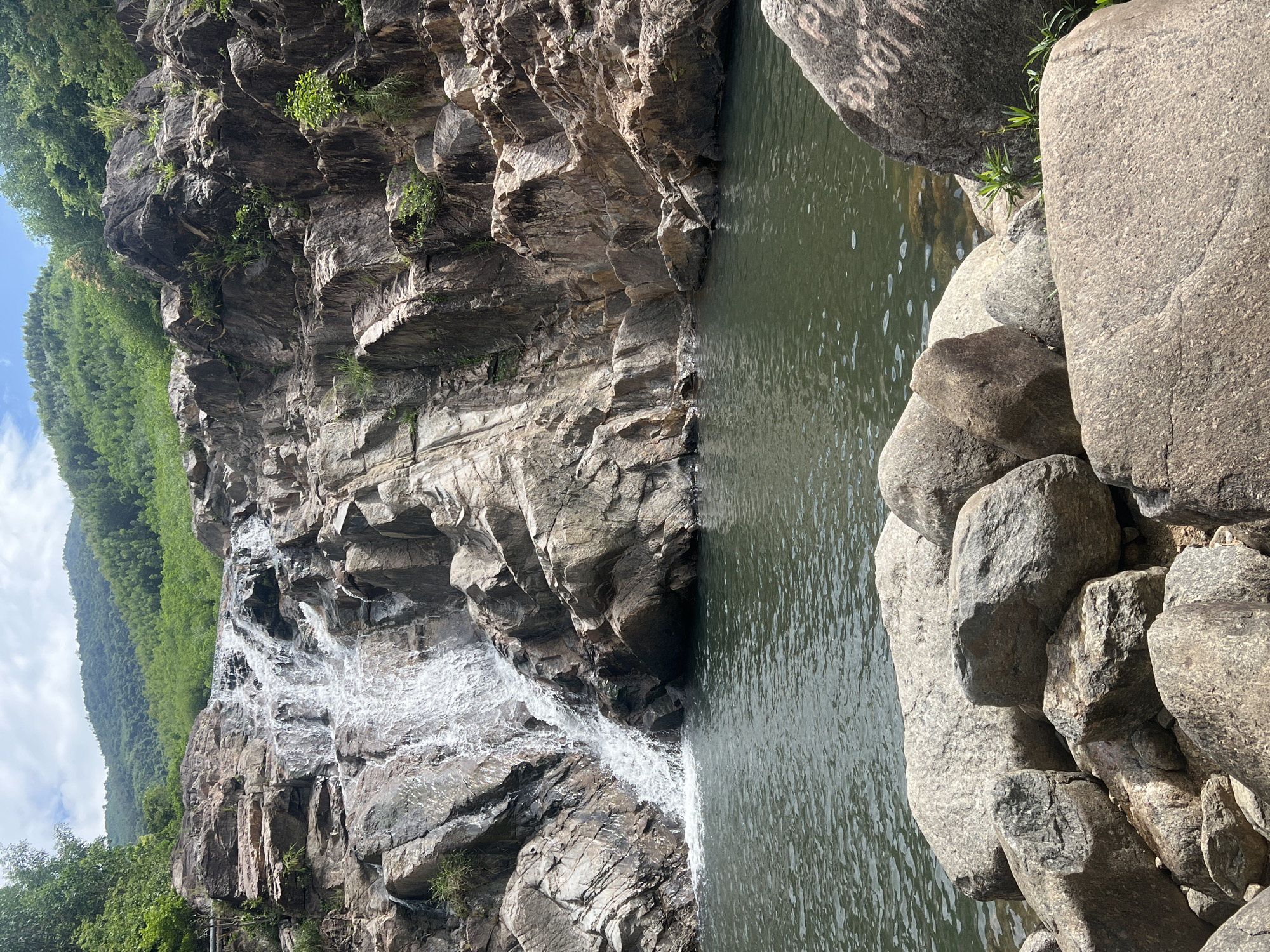 The image depicts a natural landscape featuring a cascading waterfall flowing over rugged, grey and brown rocks. The waterfall empties into a calm, dark green body of water in the foreground, which appears to be a natural pool or basin. The surrounding rocks are varied in size and texture, with some appearing smooth and others rough and jagged. Vegetation, including green trees and bushes, is visible on the slopes behind the waterfall and on the rocky outcrops. The sky above is partly cloudy with a bright, diffused light, suggesting daytime. In the foreground, several large rocks are scattered around the edge of the water. One of these rocks on the right side of the frame has faint, chalky white writing on its surface, which is difficult to decipher but appears to be text. The scene is likely a natural tourist attraction or a hiking destination, possibly in a mountainous or forested area. The writing on the rock suggests human presence and interaction with the environment. Based on the general geography and common tourist spots in Đà Nẵng, this could be a scene from one of its natural attractions like Suối Đá Falls or the waterfalls within the Marble Mountains park area, though a definitive identification without further context is not possible. The overall impression is one of natural beauty and tranquility.