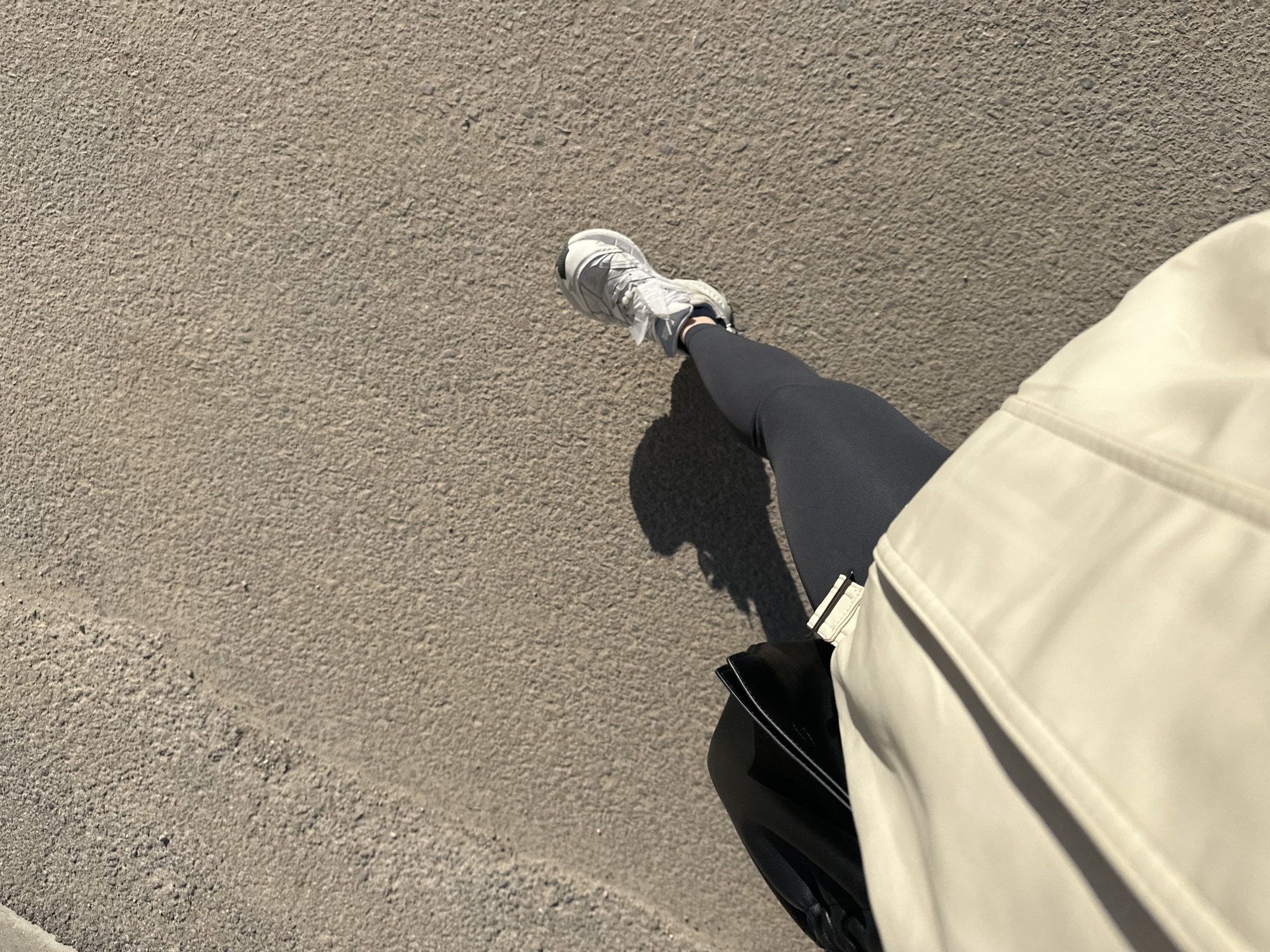 An individual is walking on a paved surface, likely a sidewalk or pedestrian area. The person is wearing a grey athletic shoe, black leggings, and a light-colored jacket or coat. A black bag is also visible near the individual's leg. The surface has a rough, textured appearance. Due to the lack of identifiable landmarks, it is impossible to determine if the location is in the city of Sofia, Bulgaria.
