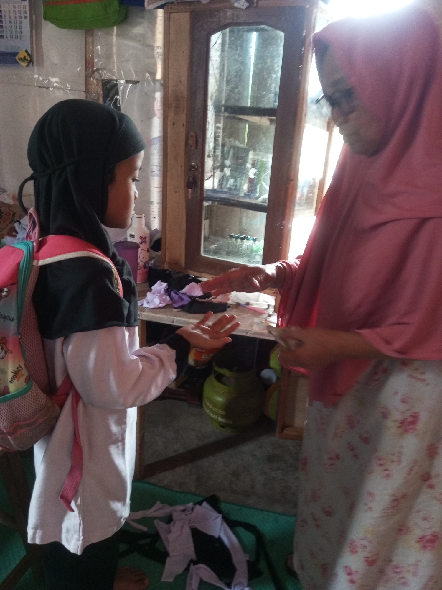 In a room in Kalibebe, Indonesia, a young girl wearing a black hijab and a white and black uniform, carrying a pink backpack, stands to the left of the frame. To her right, an older woman in a pink hijab and a floral patterned dress, wearing glasses, is handing her something. In the background, there is a wooden cabinet with glass doors, and various items are scattered on shelves and surfaces, including bottles, colorful fabric scraps, and a green gas cylinder. The floor is a mix of concrete and a green mat.