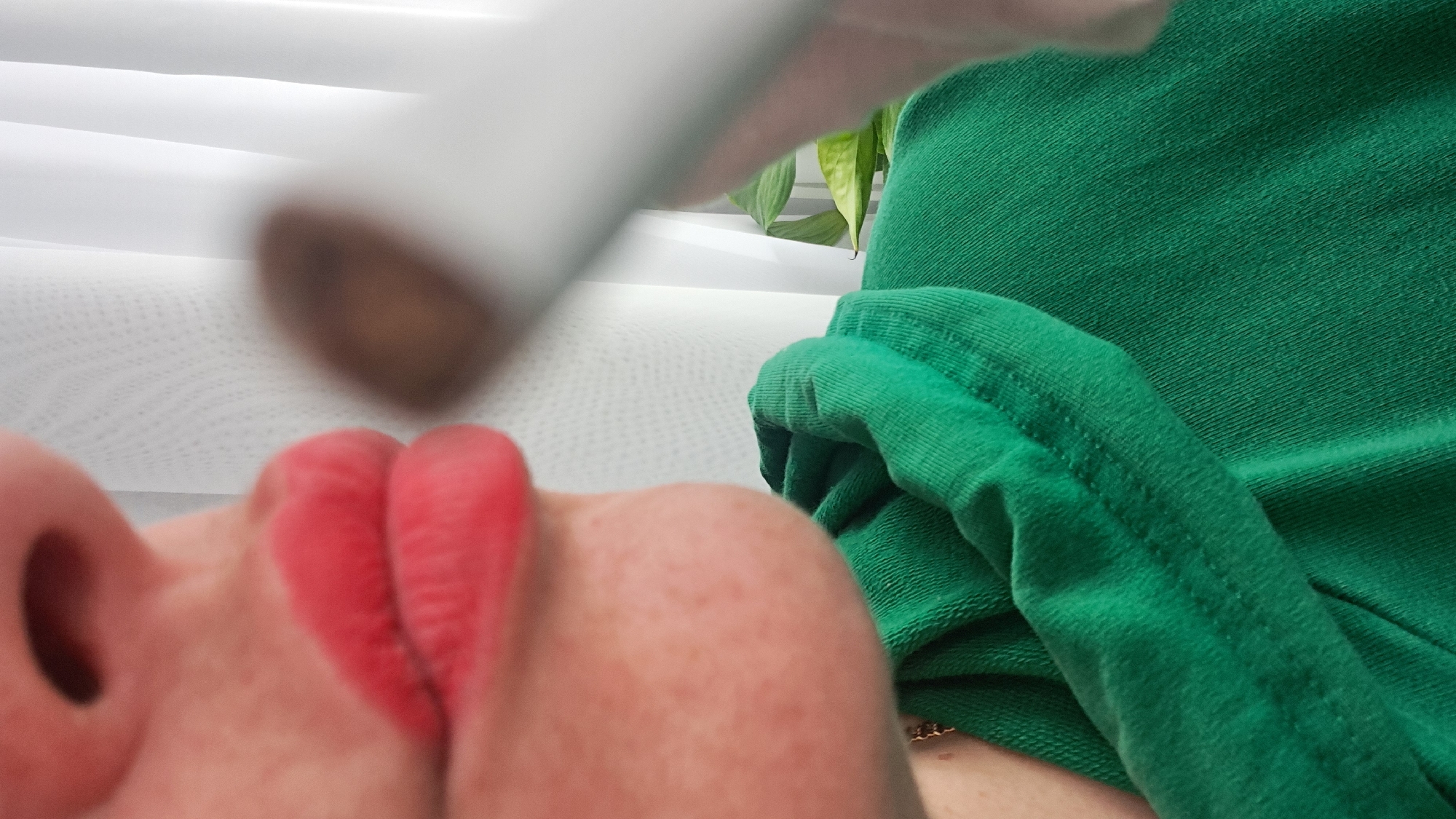 A close-up, upward-angled view focuses on a person's face and upper body. The person's face is prominent, featuring a nose and lips colored red. The skin is fair with visible freckles. To the right, a green fabric garment is seen. Above the face is a blurry, elongated white object with a brown tip. The background includes horizontal white lines, possibly blinds, and some green foliage. There is no information available to determine the location is Elektrostal, Russia.