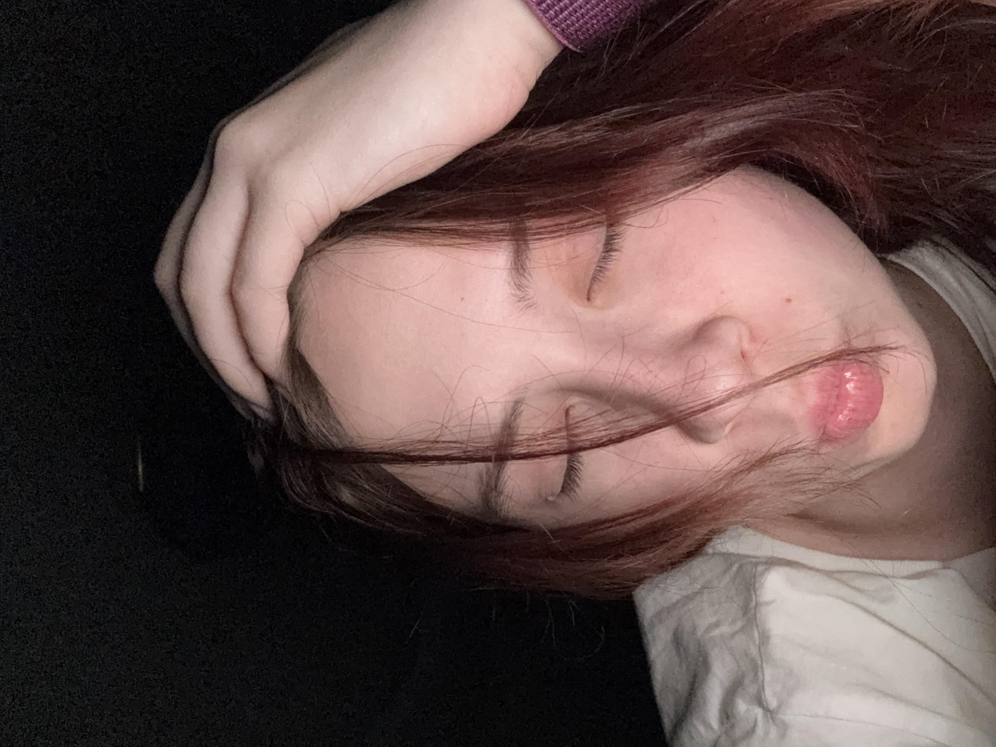 A close-up, downward-angled photograph depicts a woman's face, eyes closed, with a strand of auburn hair across her face. Her hand is resting on her head. Her skin appears smooth and pale. The background appears to be a dark, undefined area. A white garment is visible at the right edge of the frame. The setting is not identifiable, and no information relates to Belaya Pashnya, Russia.