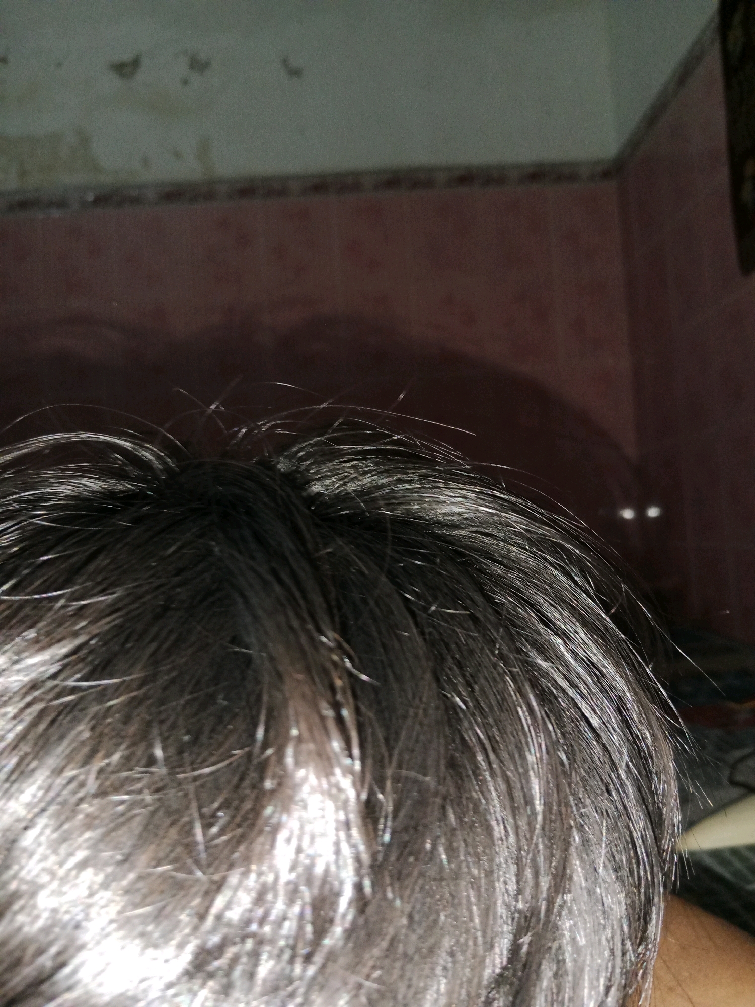 The image displays the top of a person's head, with dark, slightly damp hair in the foreground. The hair appears somewhat disheveled, with individual strands catching the light. In the background, the walls of a room are visible. One wall is a muted pale green with some discoloration and what appear to be water stains or peeling paint. Below this is a band of patterned wallpaper in shades of pink and brown. To the right, another wall is a deep pink or reddish-brown. Notably, two small, bright white points of light, resembling eyes, are visible in the shadowy background on the pink wall. The overall lighting is dim, contributing to the shadowy effect and the reflective quality of the hair. The scene appears to be an indoor environment, possibly a residential room, and given the mention of Ringinrejo, Indonesia, it is likely a domestic setting within that location.