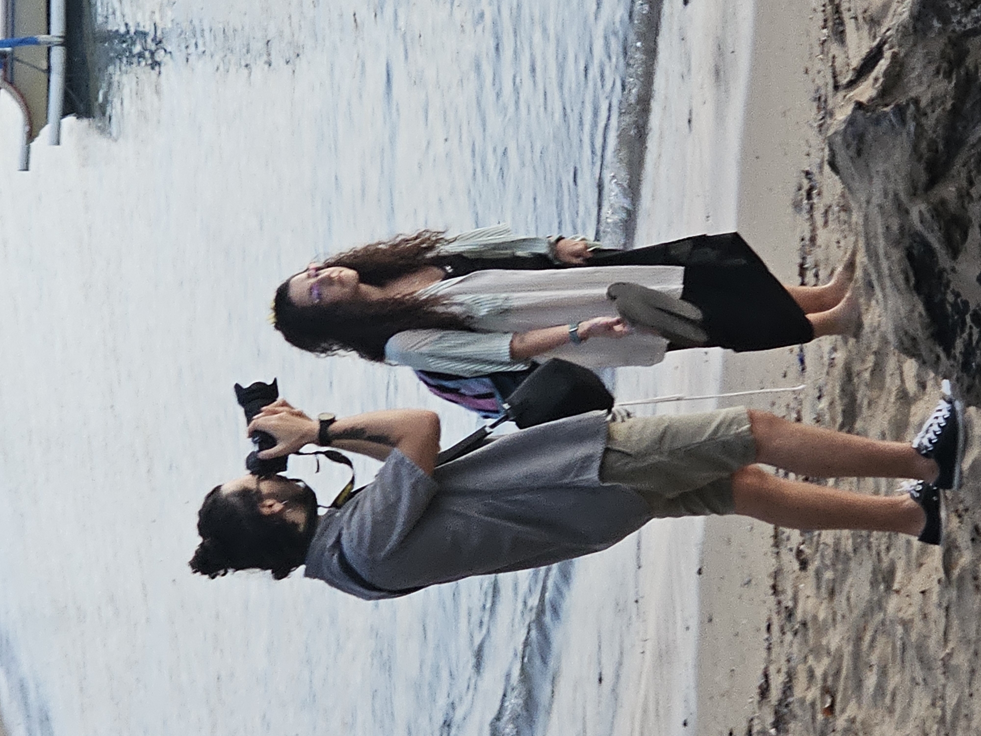 An unidentifiable person with a bun of dark hair is taking a picture with a professional camera near the water's edge on a beach. The person is wearing a gray short-sleeved button-down shirt and khaki shorts, and black Converse sneakers. Behind this person, a woman with long, dark curly hair is standing and looking out at the water. She is wearing a white cardigan over a black dress and pink-rimmed glasses. She is holding a pair of gray flip-flops in her right hand. The beach is made of sand, and small waves are coming in from the ocean. In the background, a small boat is visible on the water. The scene takes place on a beach in Denpasar, Indonesia, a city known for its beaches and vibrant culture.