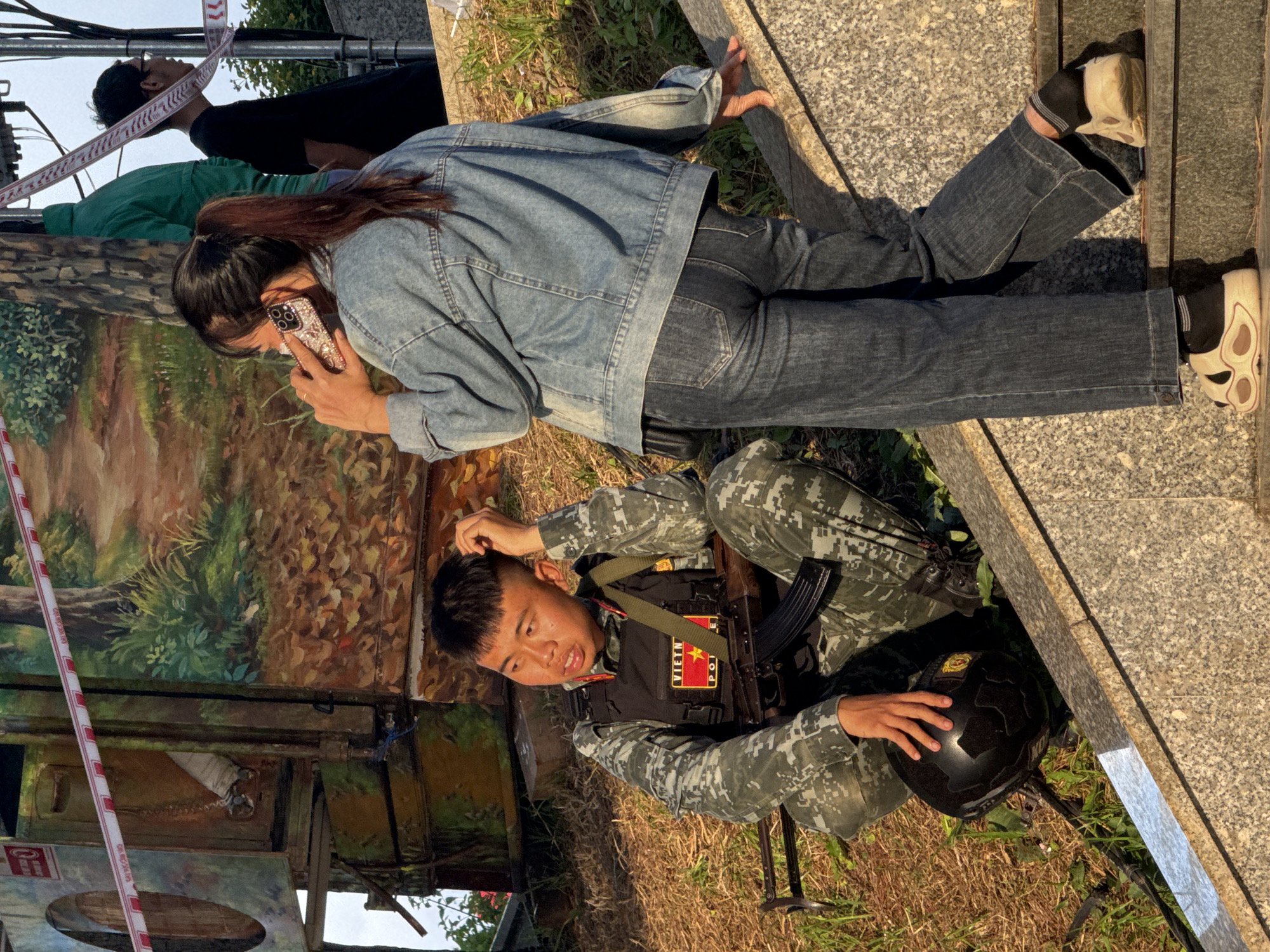 A young Vietnamese soldier in camouflage uniform, wearing a tactical vest with a "VIETNAM POLICE" patch, sits on the ground holding a black helmet and a rifle. Next to him, a young Vietnamese woman in a denim jacket and jeans talks on a smartphone. They are near a colorful, mural-decorated structure with red and white caution tape. The scene appears to be on a grassy slope with concrete steps, suggesting an outdoor public area. The lighting indicates it is late afternoon, casting long shadows. This location is identified as the Dinh 1 National Tourism Site in Da Lat, Vietnam.