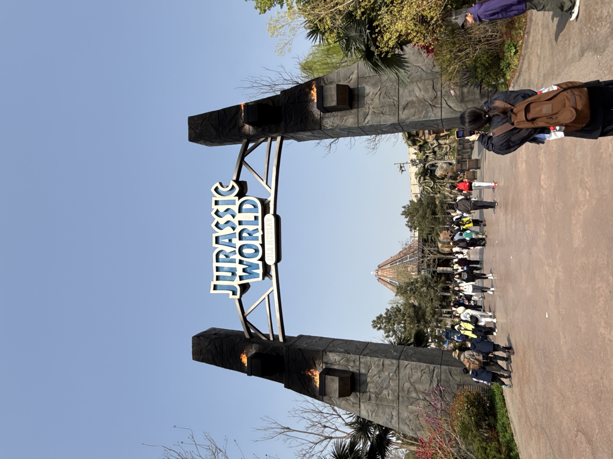 The image shows the entrance to "Jurassic World" at Universal Beijing Resort in Tongzhou District, China. The iconic archway displays the park's name in large, blue, three-dimensional letters. Flames emanate from dark, stone-like structures that frame the arch. A crowd of people is present, some walking into the park, others taking pictures. A woman with a brown backpack and black jacket is capturing a photograph of the entrance with her phone. The background reveals trees, shrubs, and part of a building. The sky is clear and blue.