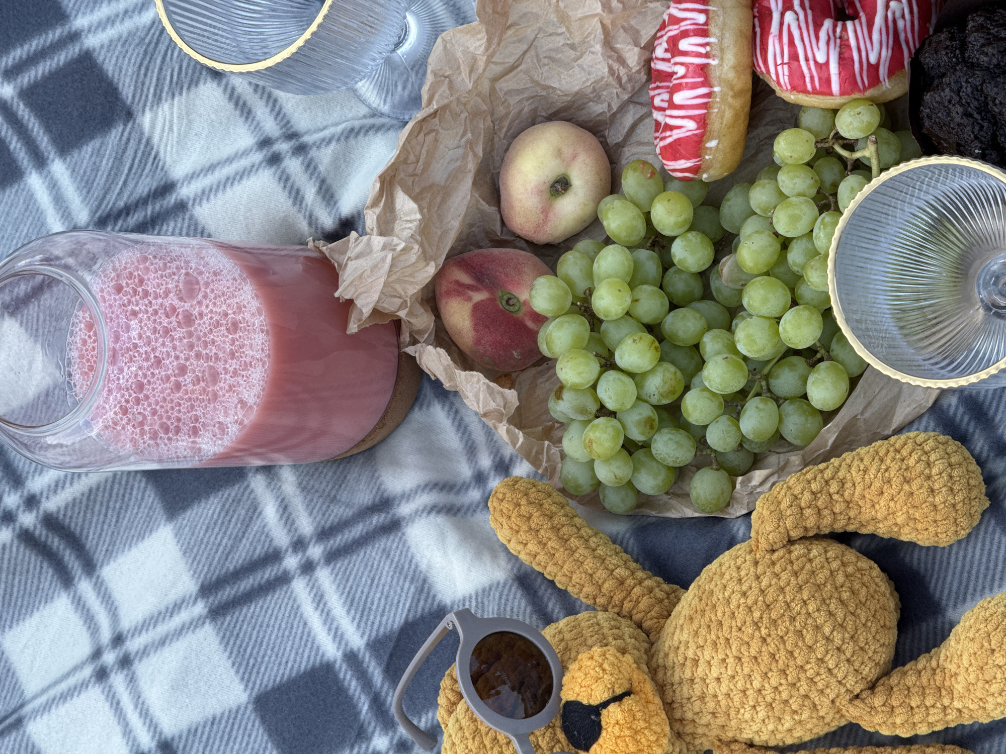 The image depicts a picnic spread on a grey and white plaid blanket. The scene includes a clear glass bottle filled with a pink, frothy beverage, a glass cup with a gold rim, a bunch of green grapes, two peaches, two donuts with pink frosting and white icing, a brown paper bag, and a yellow crocheted teddy bear wearing sunglasses. The teddy bear is positioned on the left side of the frame, with its arm extended. The grapes, peaches, and donuts are clustered together in the center and right side of the image, partially spilling out of the paper bag. The overall setting suggests a casual outdoor gathering. There is no identifiable information in the image that would place it in the city of Chaikovsky, Russia.